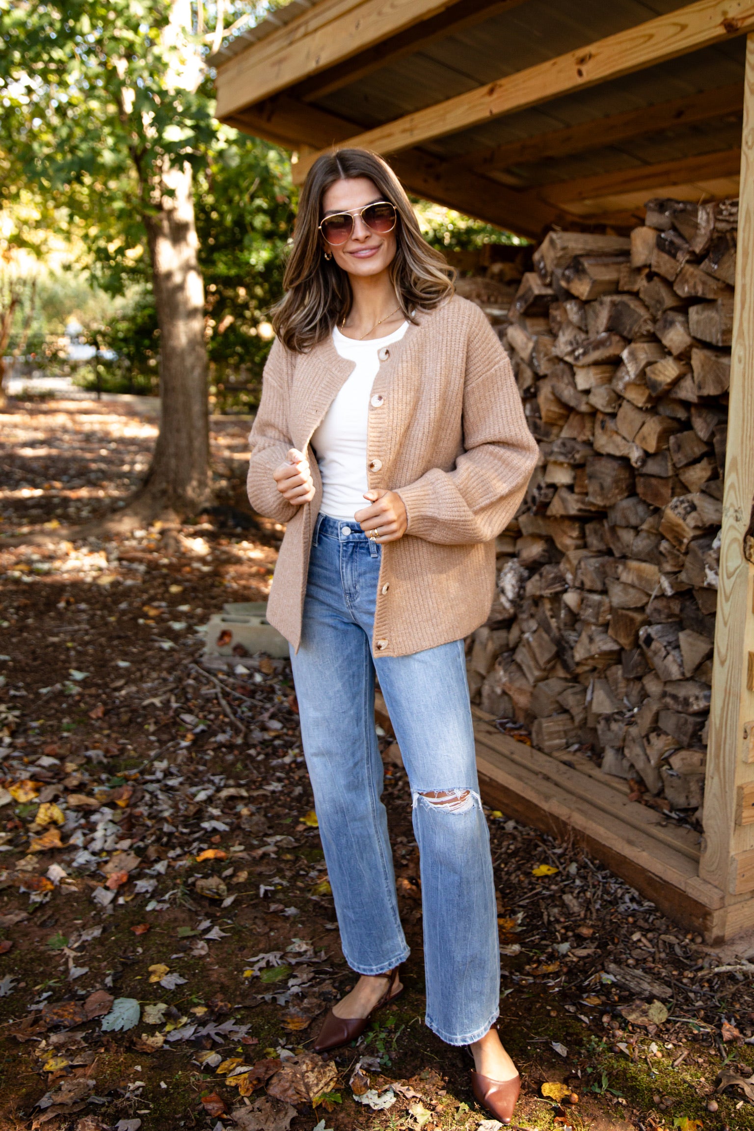Woman standing outdoors near a stack of firewood, wearing a beige cardigan, white shirt, blue jeans, and sunglasses.