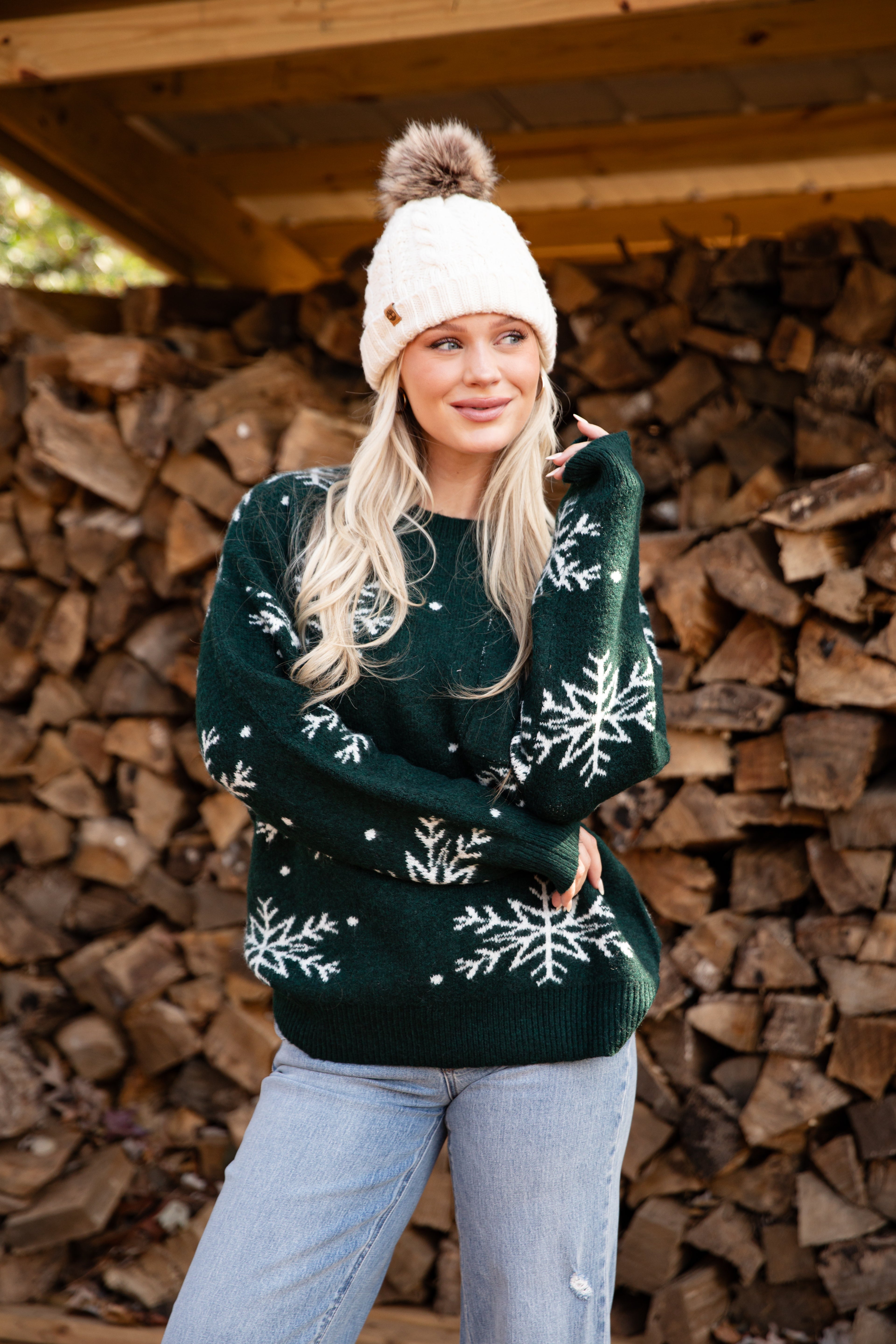 Woman wearing a green sweater with snowflake patterns and a white beanie in front of stacked firewood.