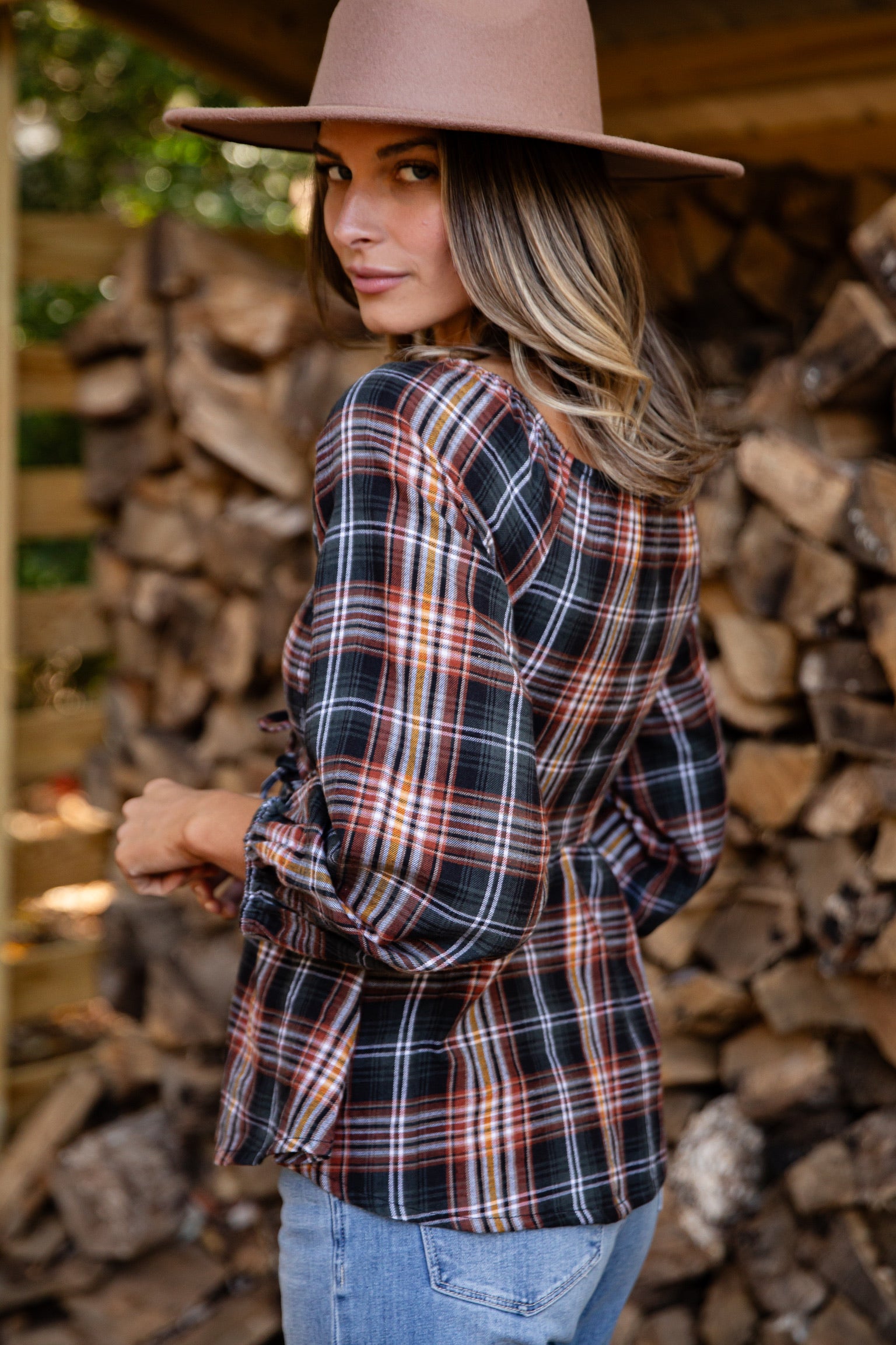 Woman wearing a plaid shirt and brown hat standing in front of stacked firewood.