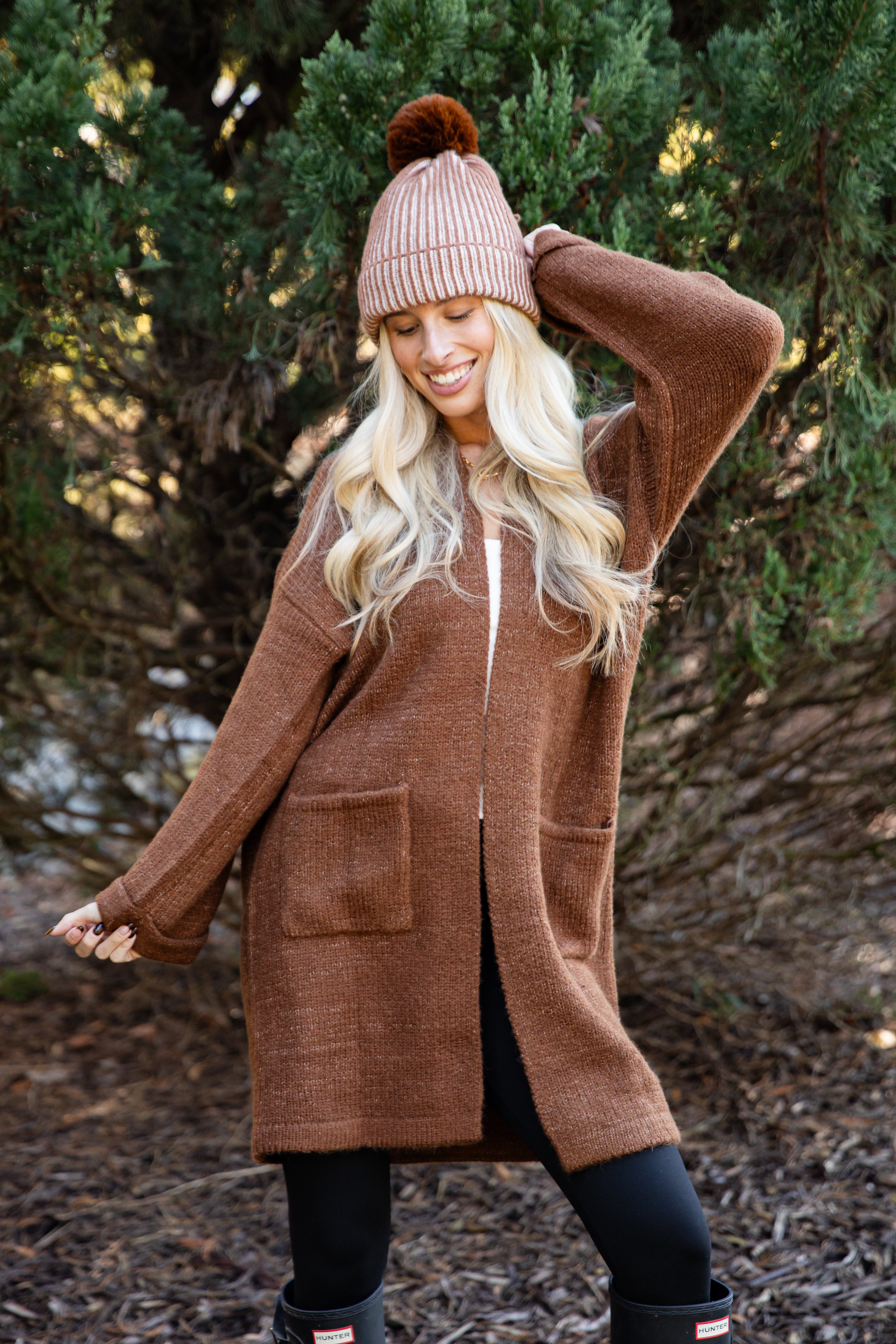Person wearing a brown cardigan, beige beanie with a pom-pom, and black boots in a forest setting.
