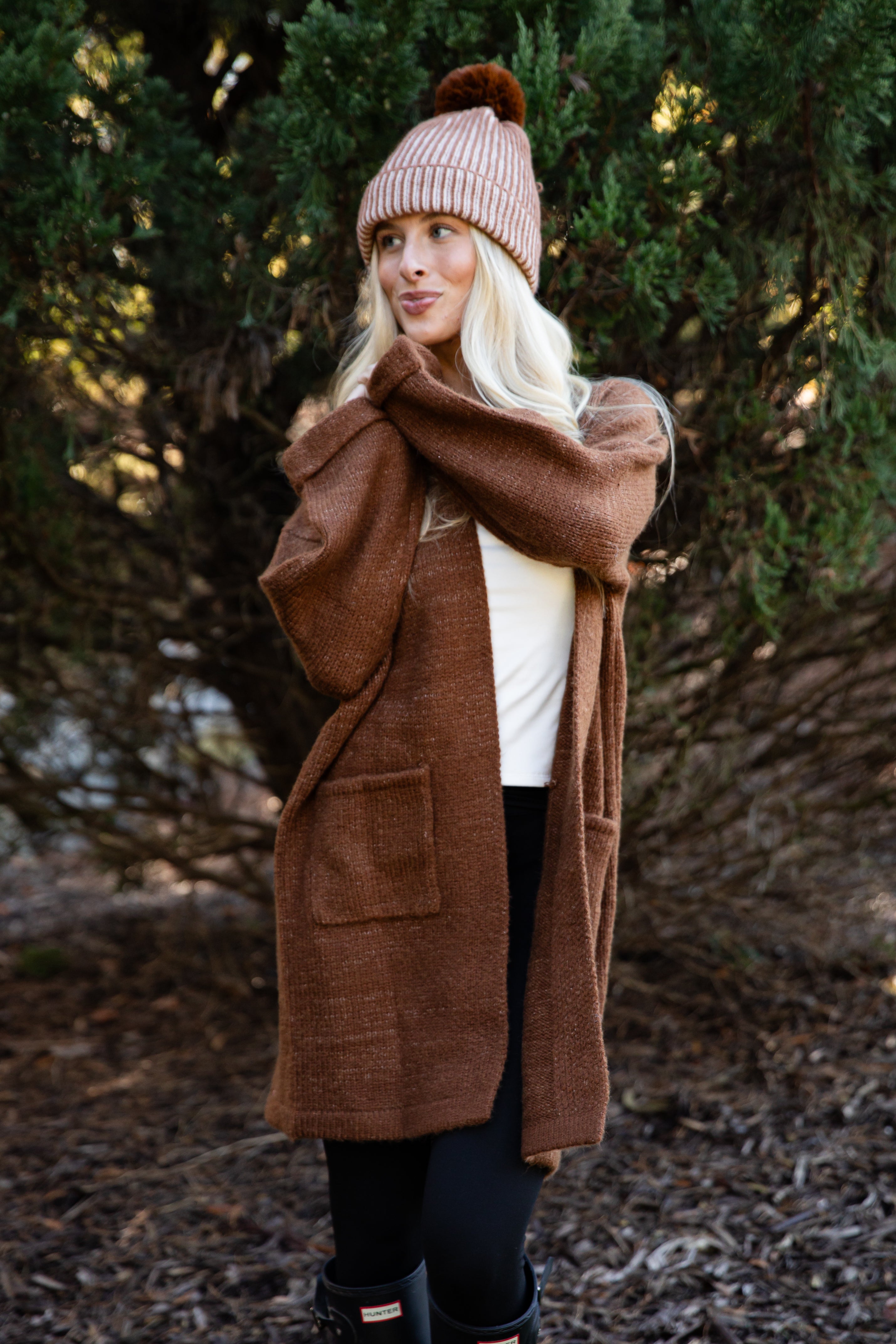 Woman wearing a brown coat and pink beanie standing in a forest setting
