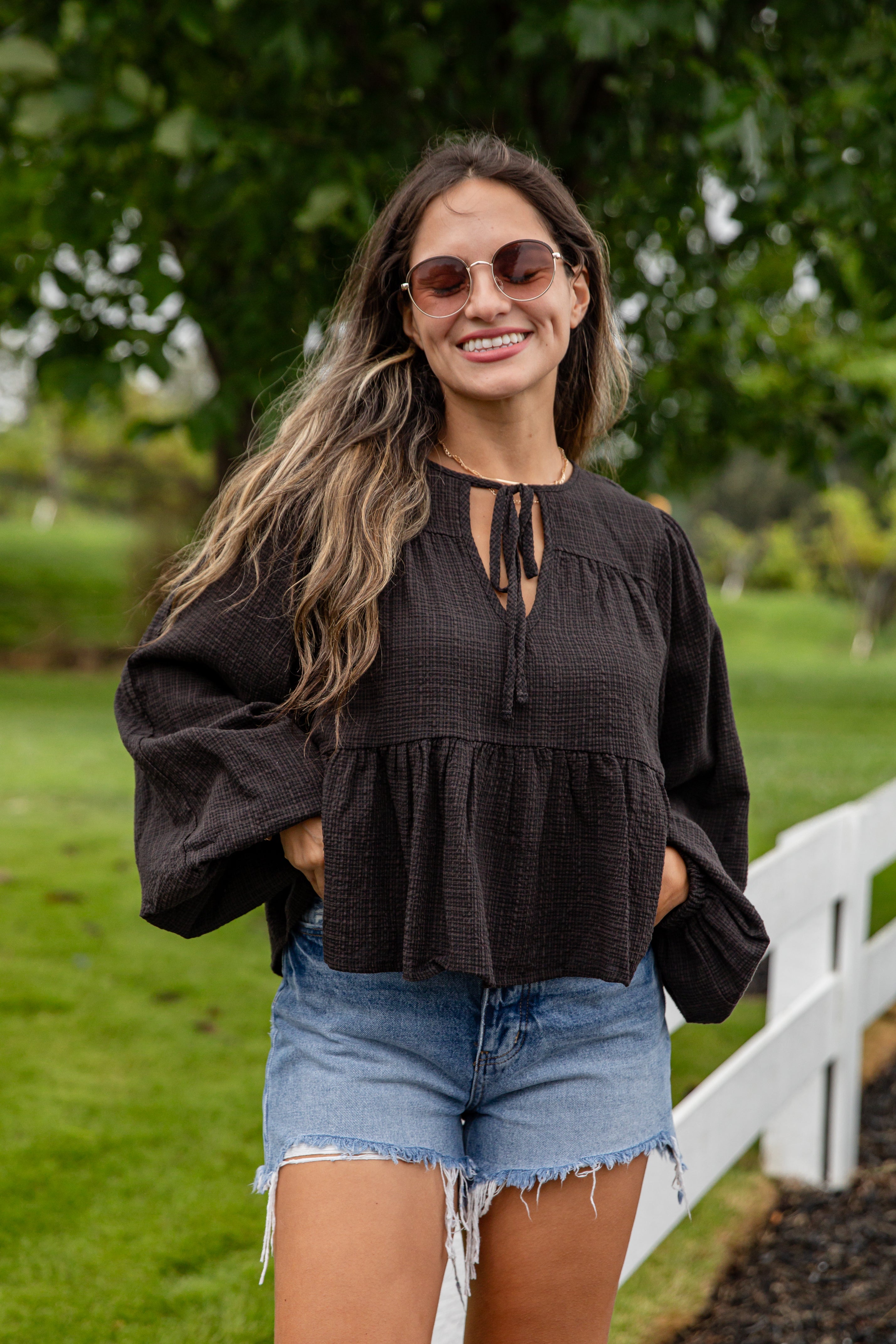 Woman wearing a black top and denim shorts standing outdoors with greenery in the background