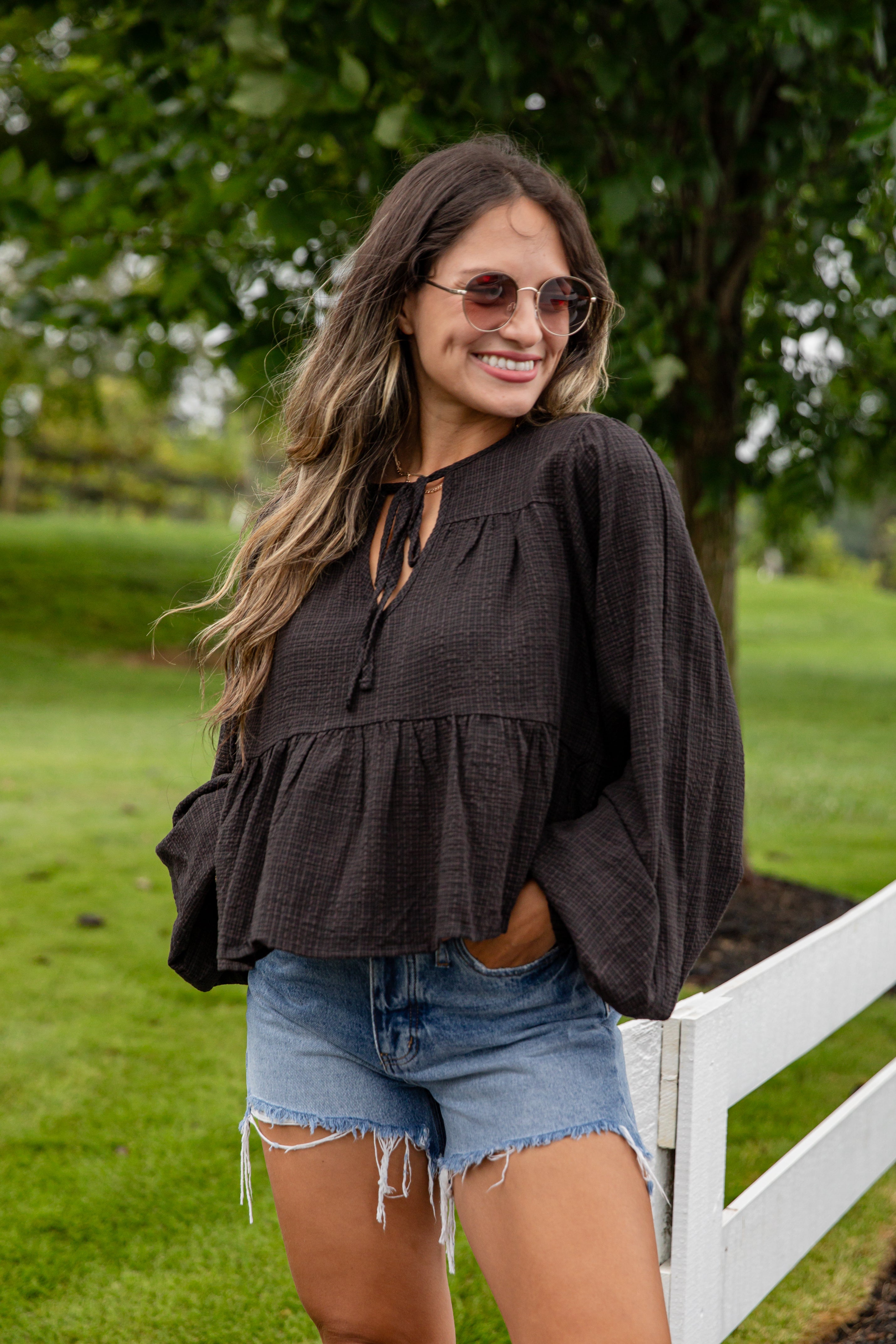 Woman wearing a black top and denim shorts standing outdoors with trees in the background