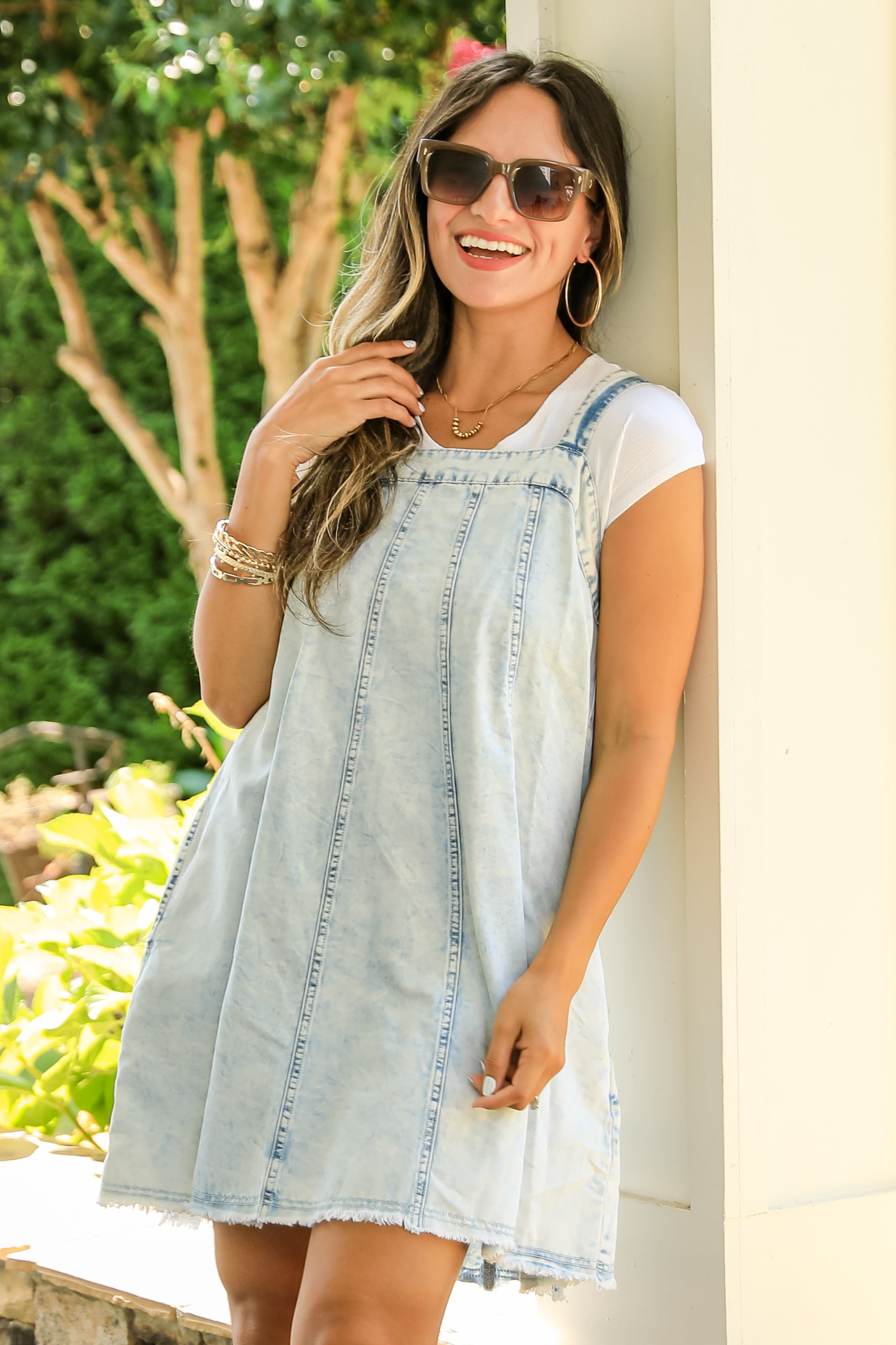 Woman wearing a light blue denim dress and sunglasses, standing against a white wall with greenery in the background.