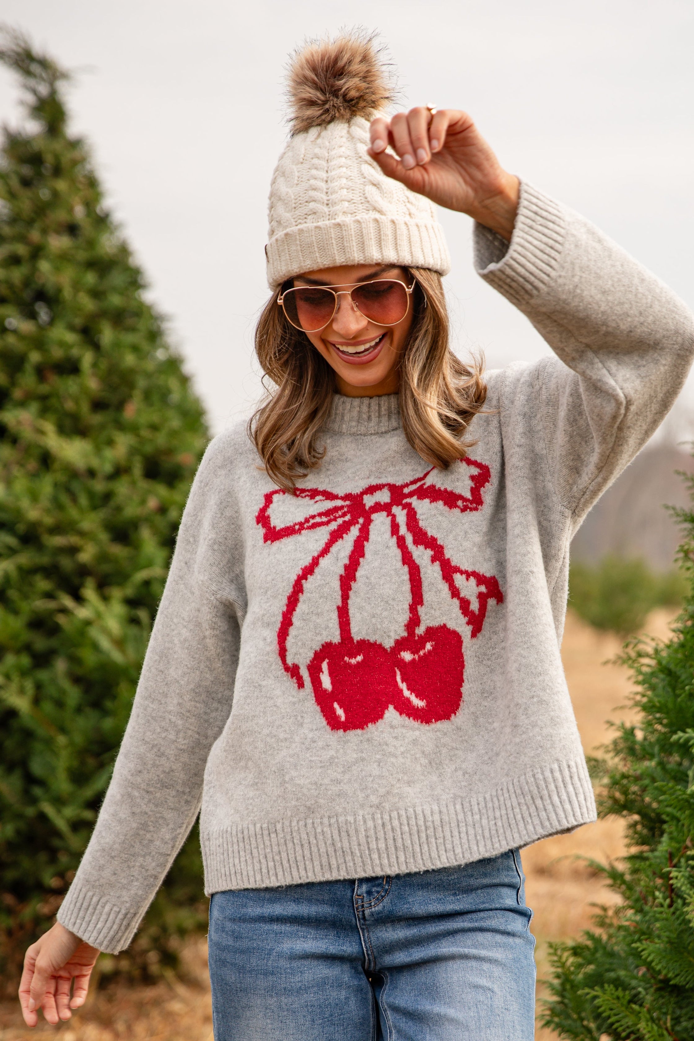 Woman wearing a gray sweater with red cherry design, white beanie, and sunglasses outdoors.