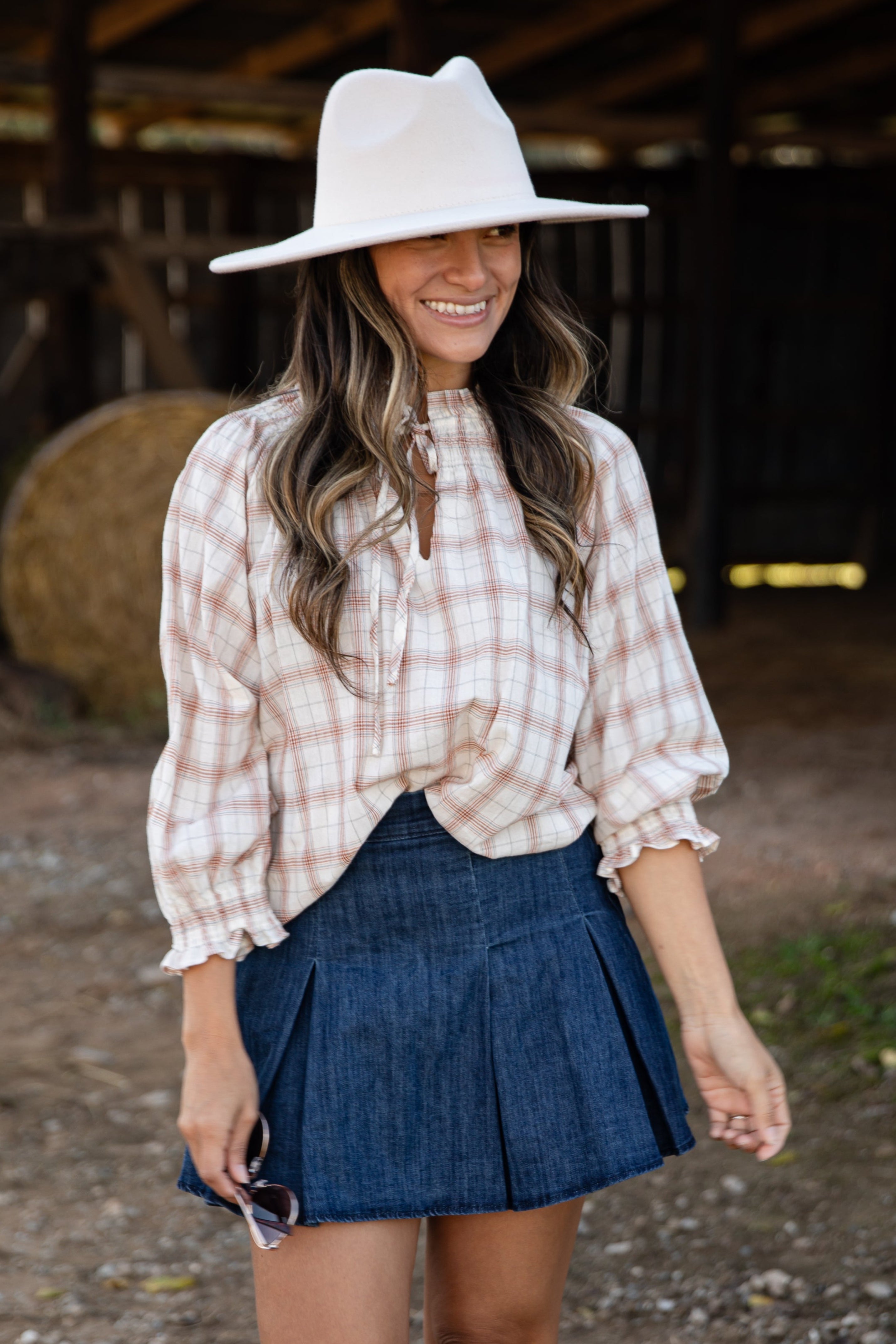 Woman wearing a plaid shirt, denim skirt, and cowboy boots in a barn setting.