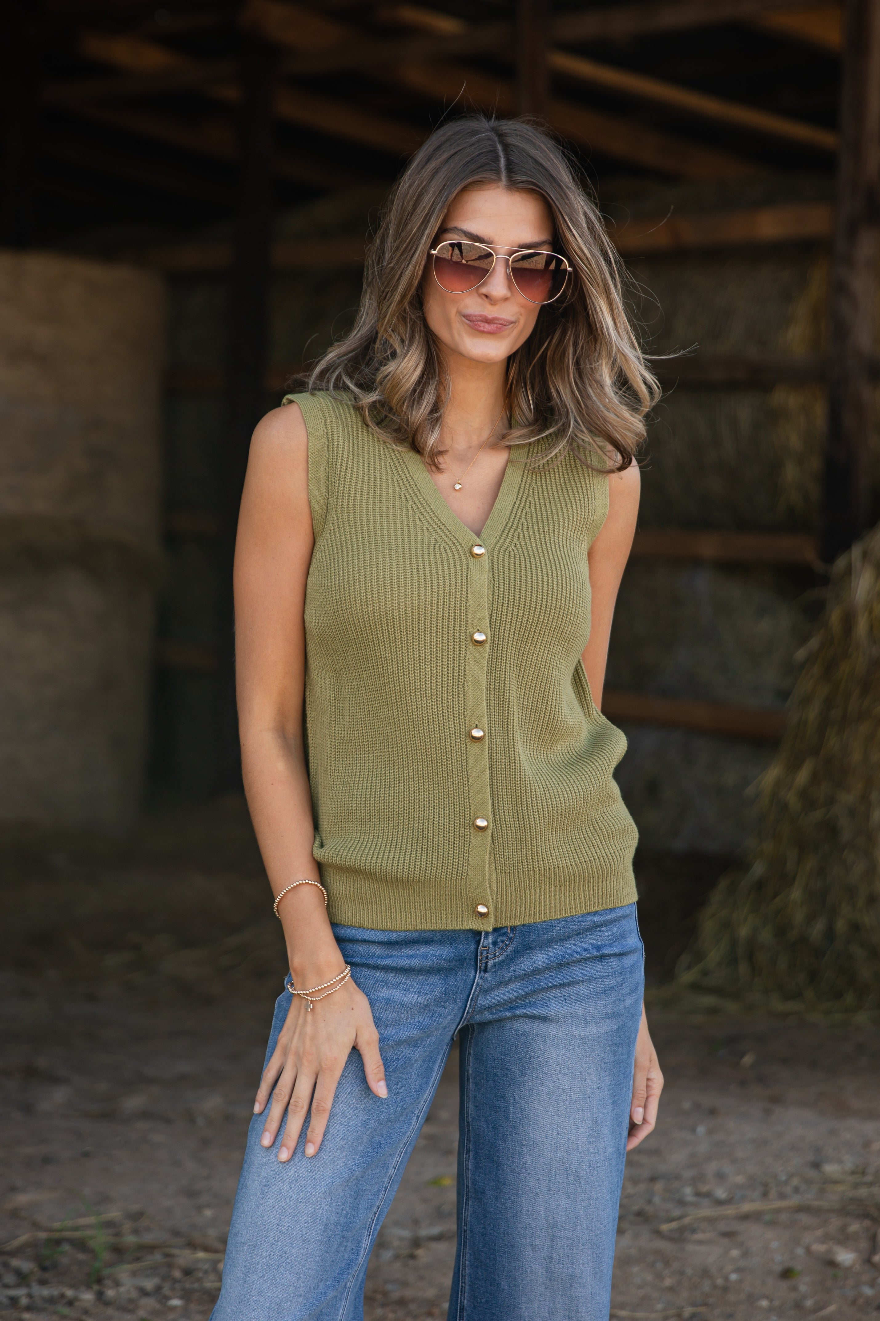Woman wearing a green sleeveless sweater and blue jeans in a rustic setting
