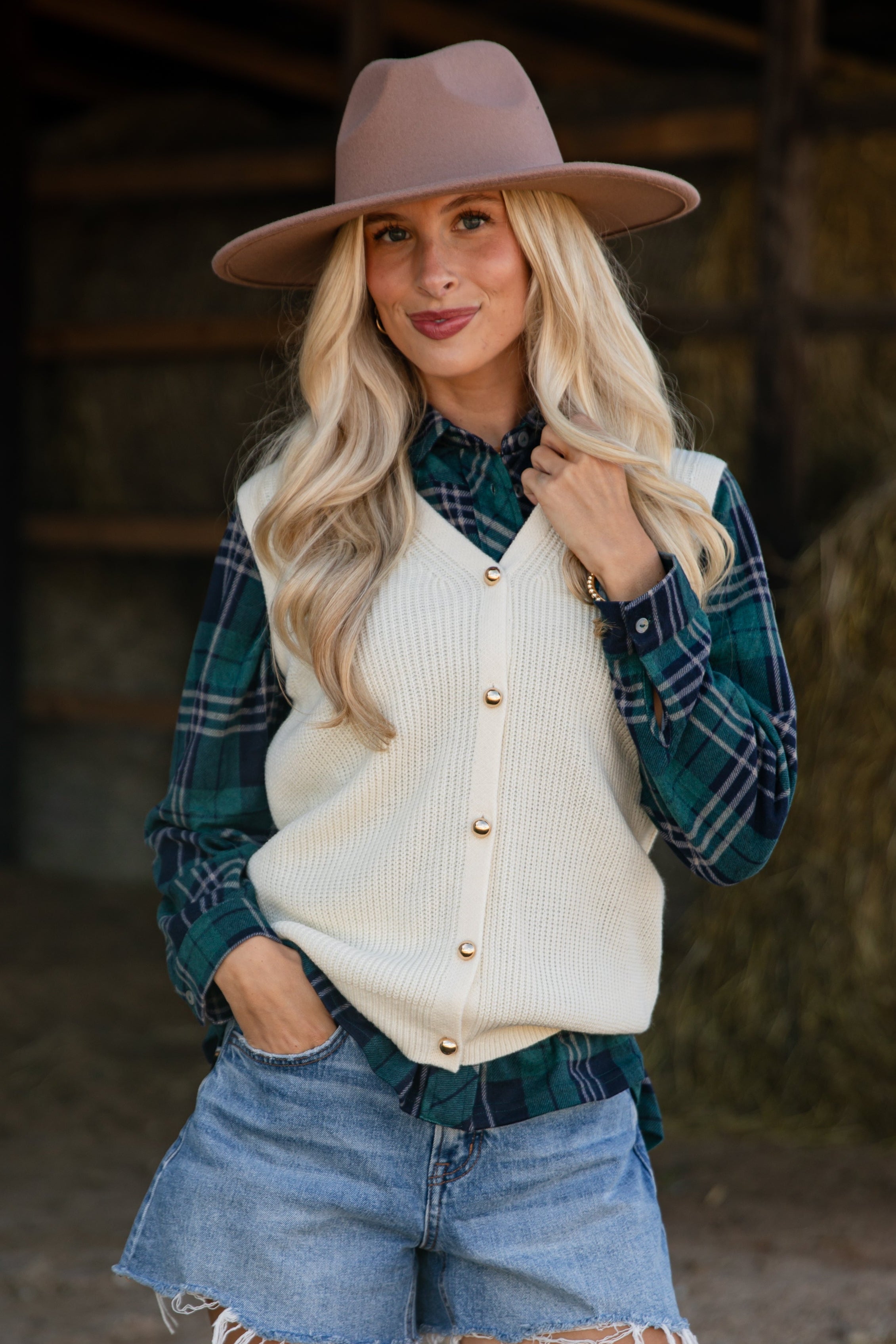Woman wearing a beige hat, plaid shirt, cream cardigan, denim shorts, and white sneakers in a barn setting.