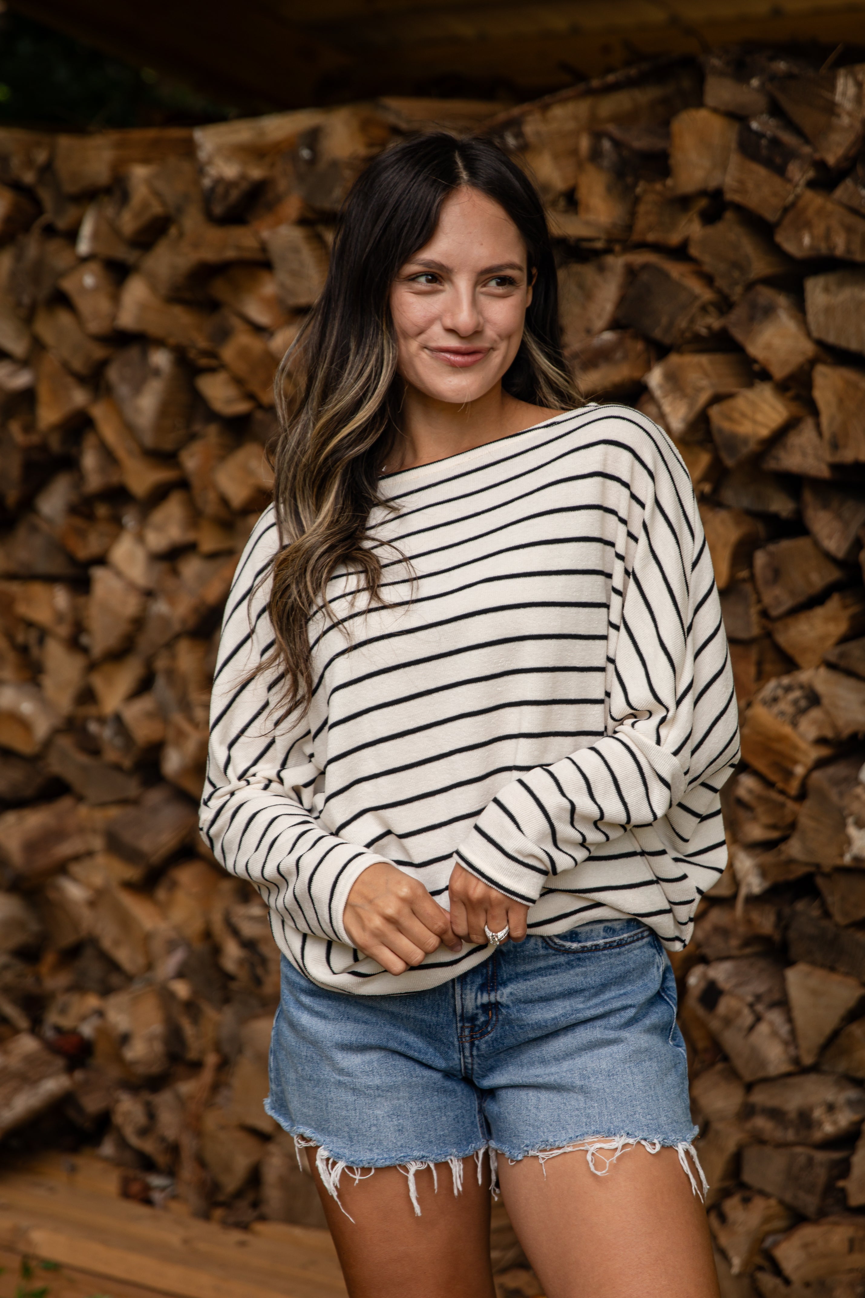 Woman wearing a striped sweater and denim shorts standing in front of stacked firewood.
