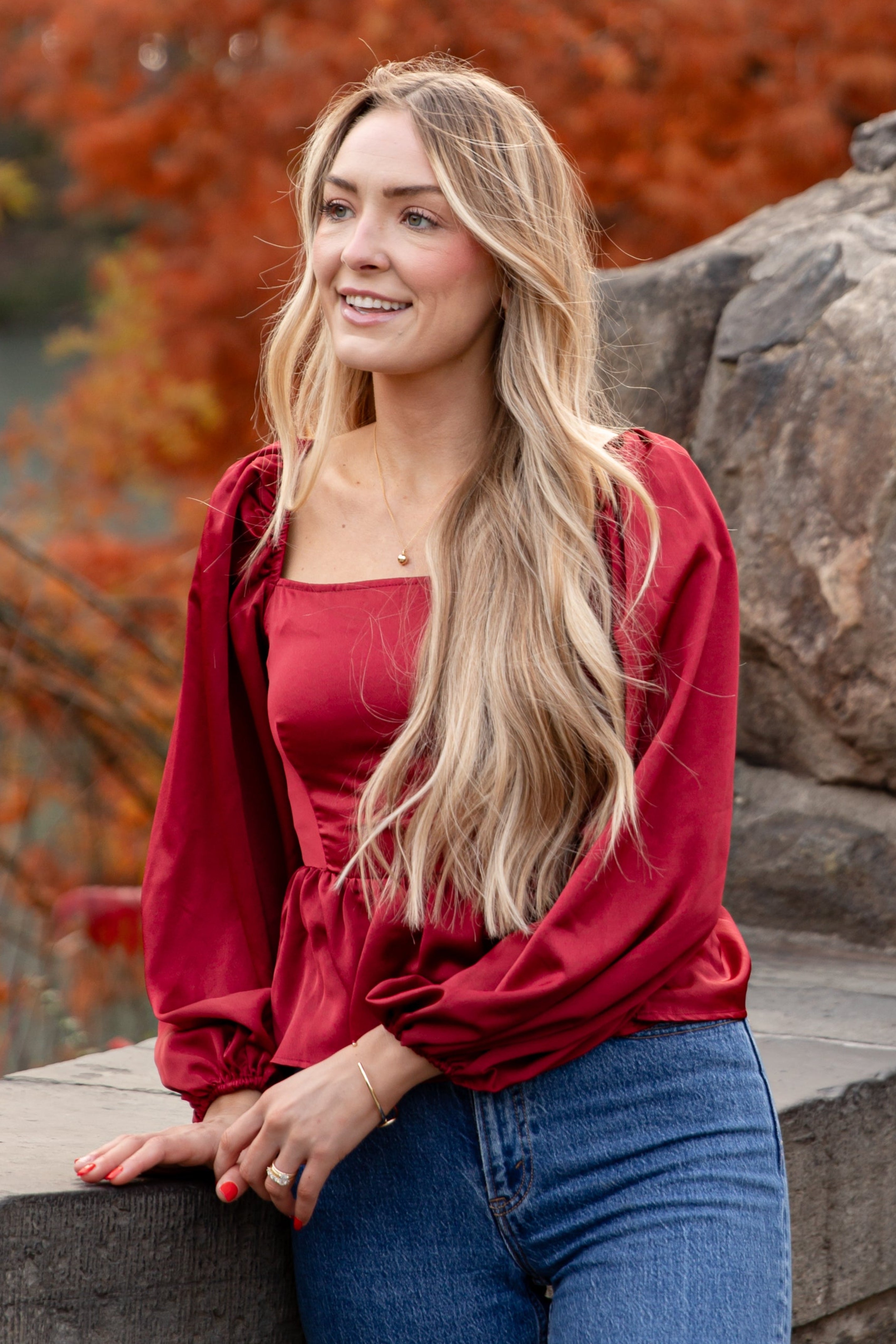 Woman in a red top and blue jeans leaning against a stone wall with autumn trees in the background
