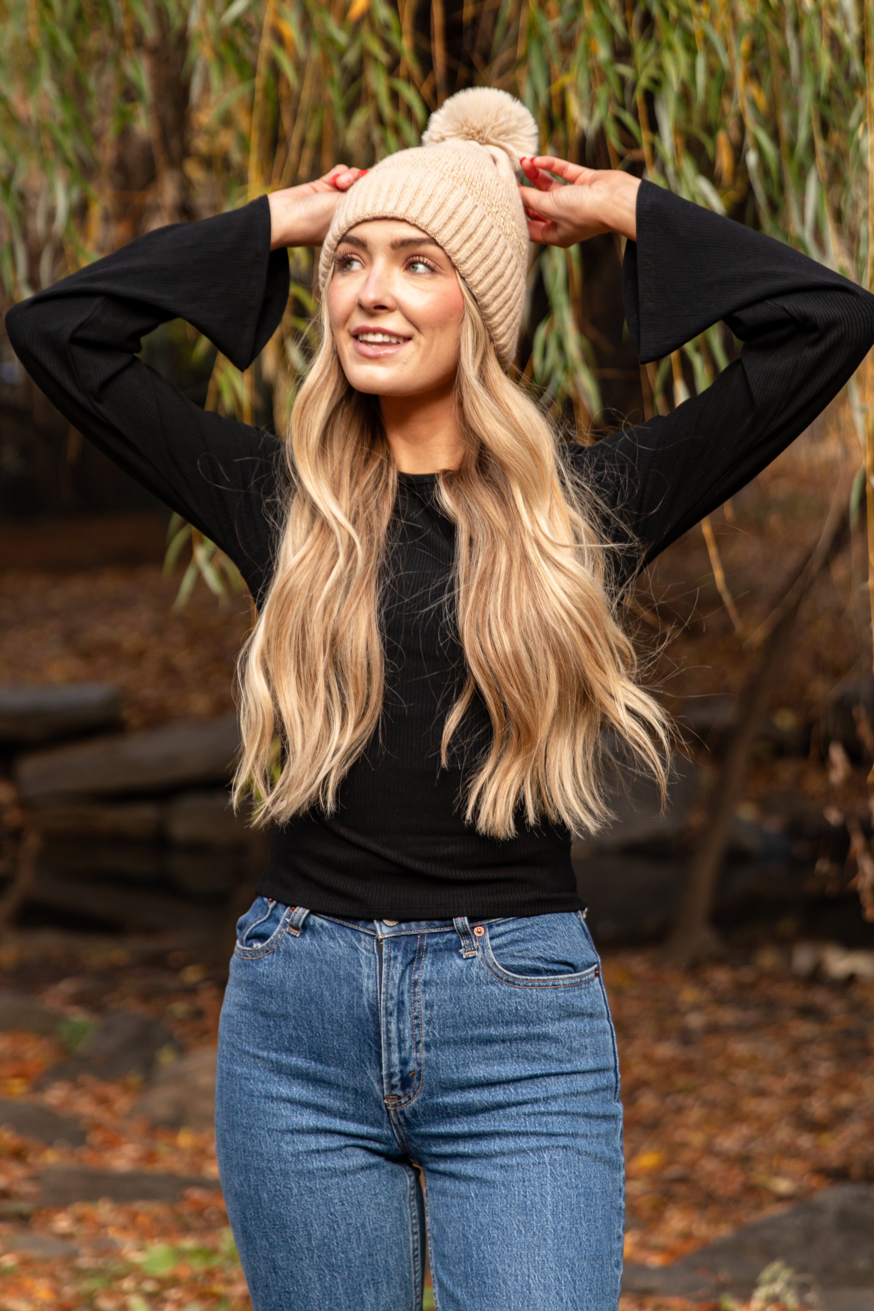 Woman wearing a beige beanie and black sweater in an outdoor setting with greenery.