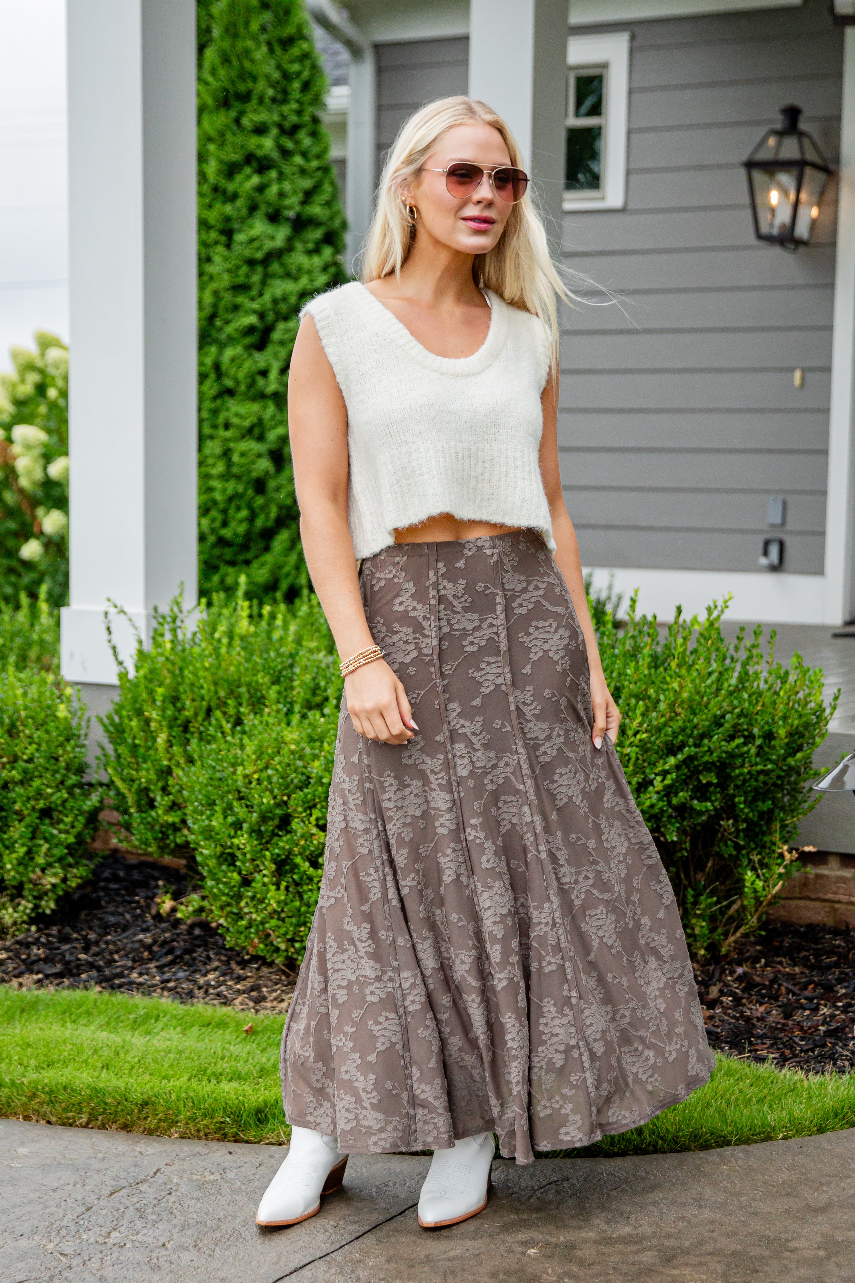 Woman wearing a white sleeveless top and brown patterned skirt standing outside a house.