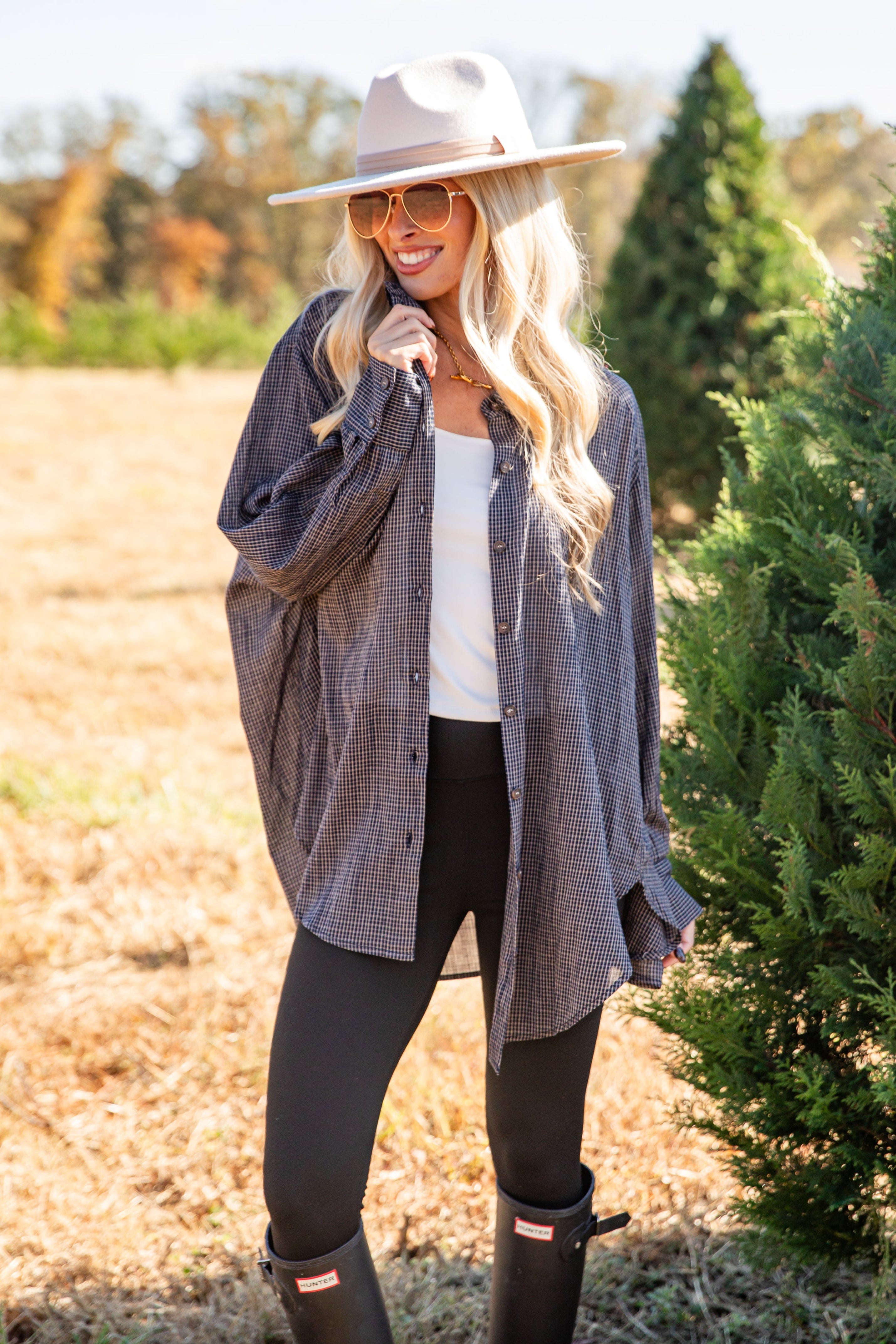 Woman standing outdoors in a field with trees, wearing a plaid shirt, white top, black pants, and boots.