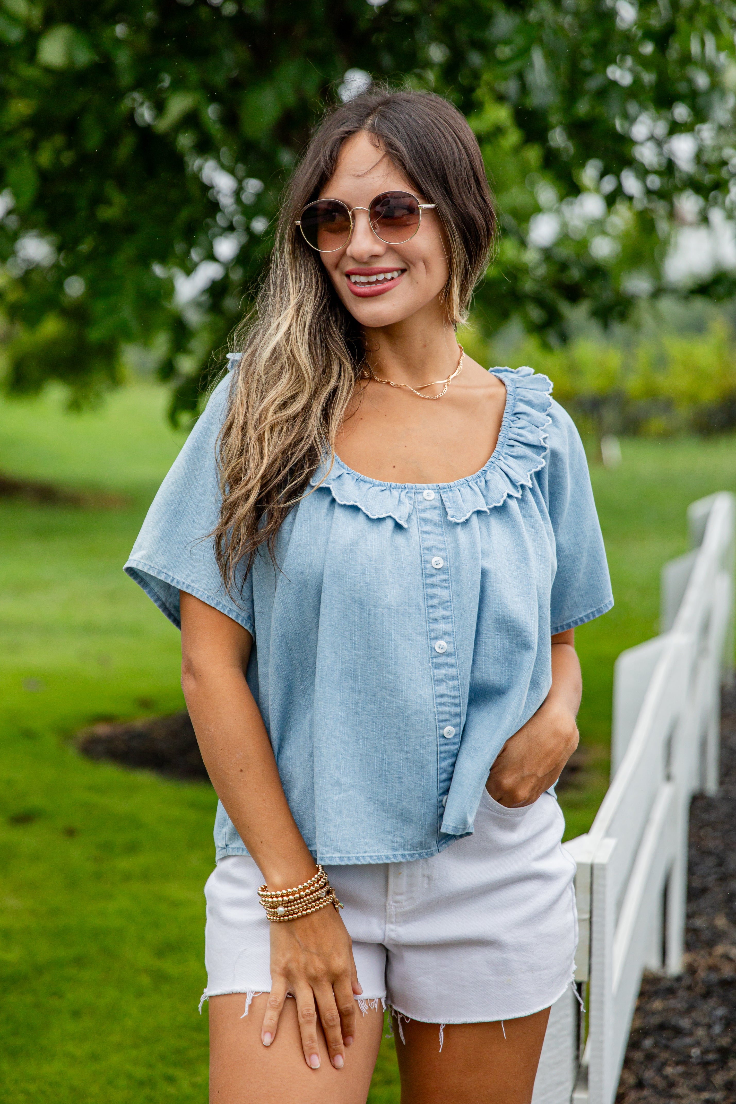 Woman wearing a light blue blouse with ruffled details and white shorts, standing outdoors.