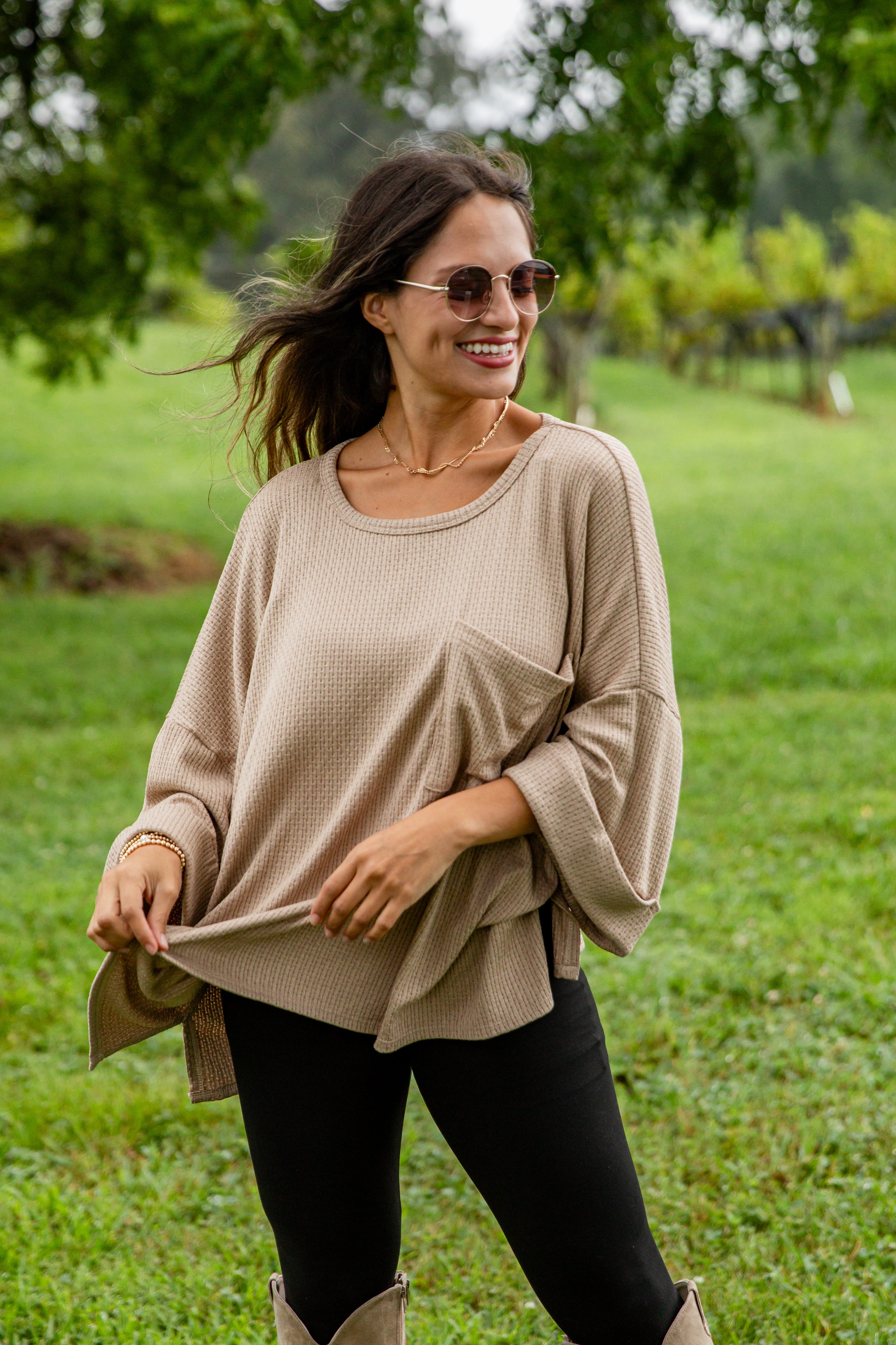 Woman in a beige top and black pants standing in a grassy field with trees in the background