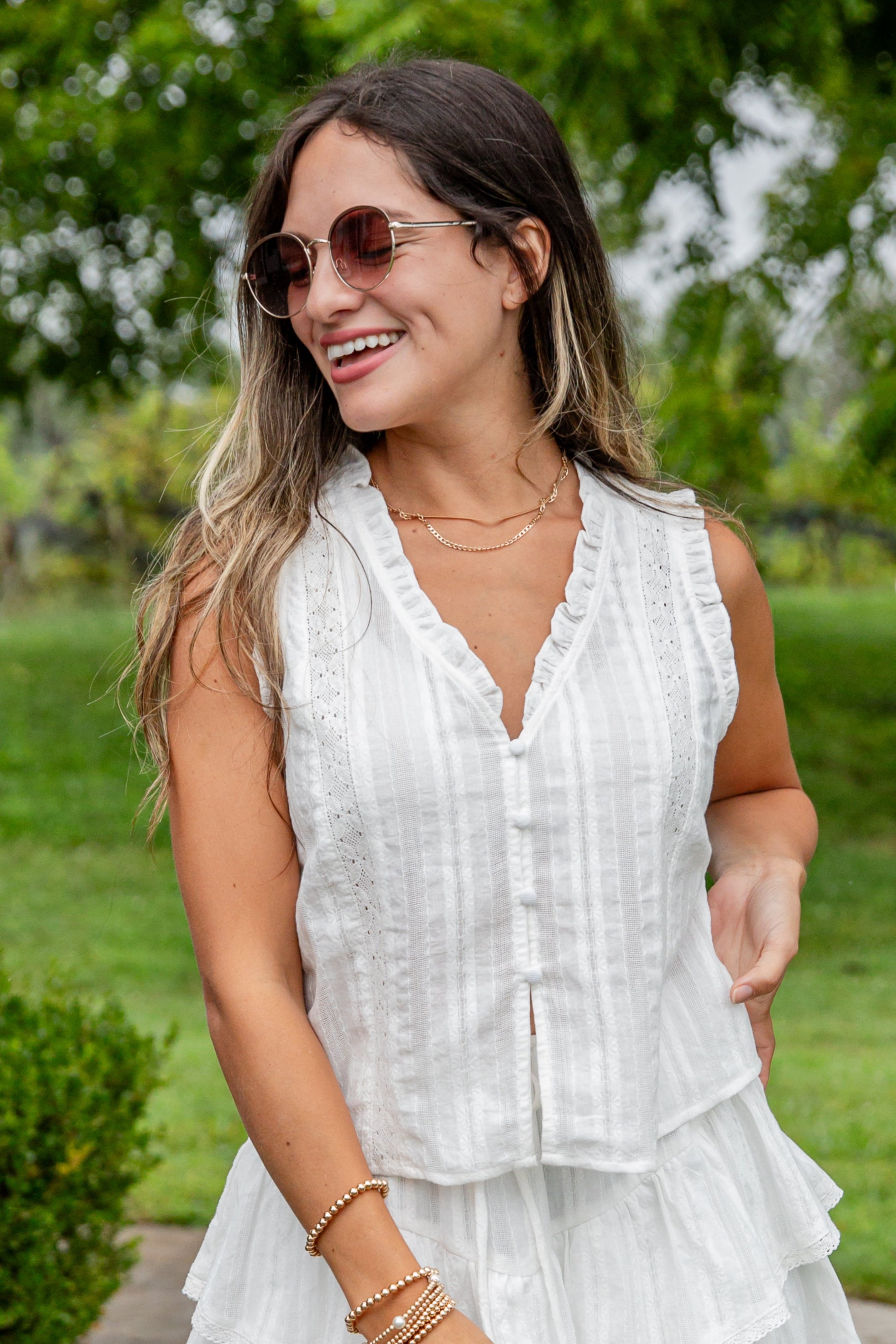 Woman in a white sleeveless dress standing outdoors with greenery in the background