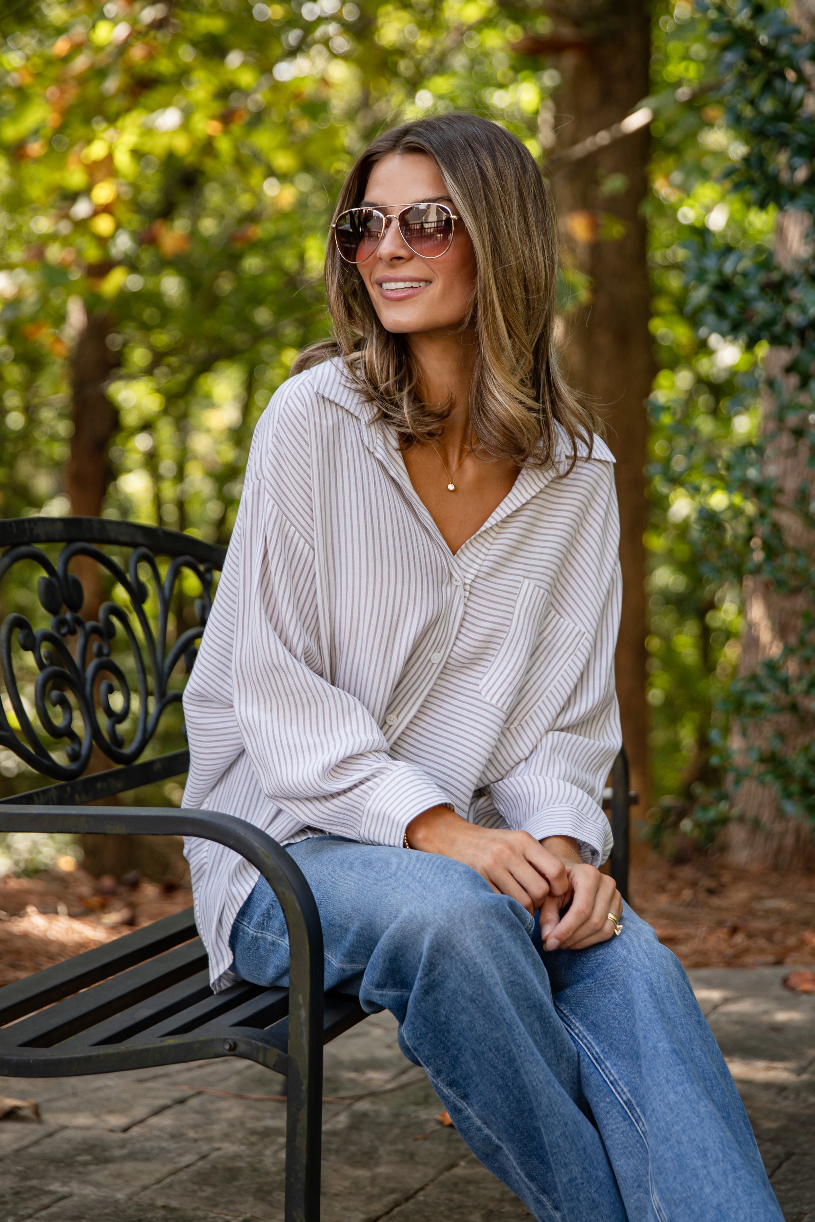 Woman sitting on a bench outdoors wearing sunglasses and a light-colored top with blue jeans.
