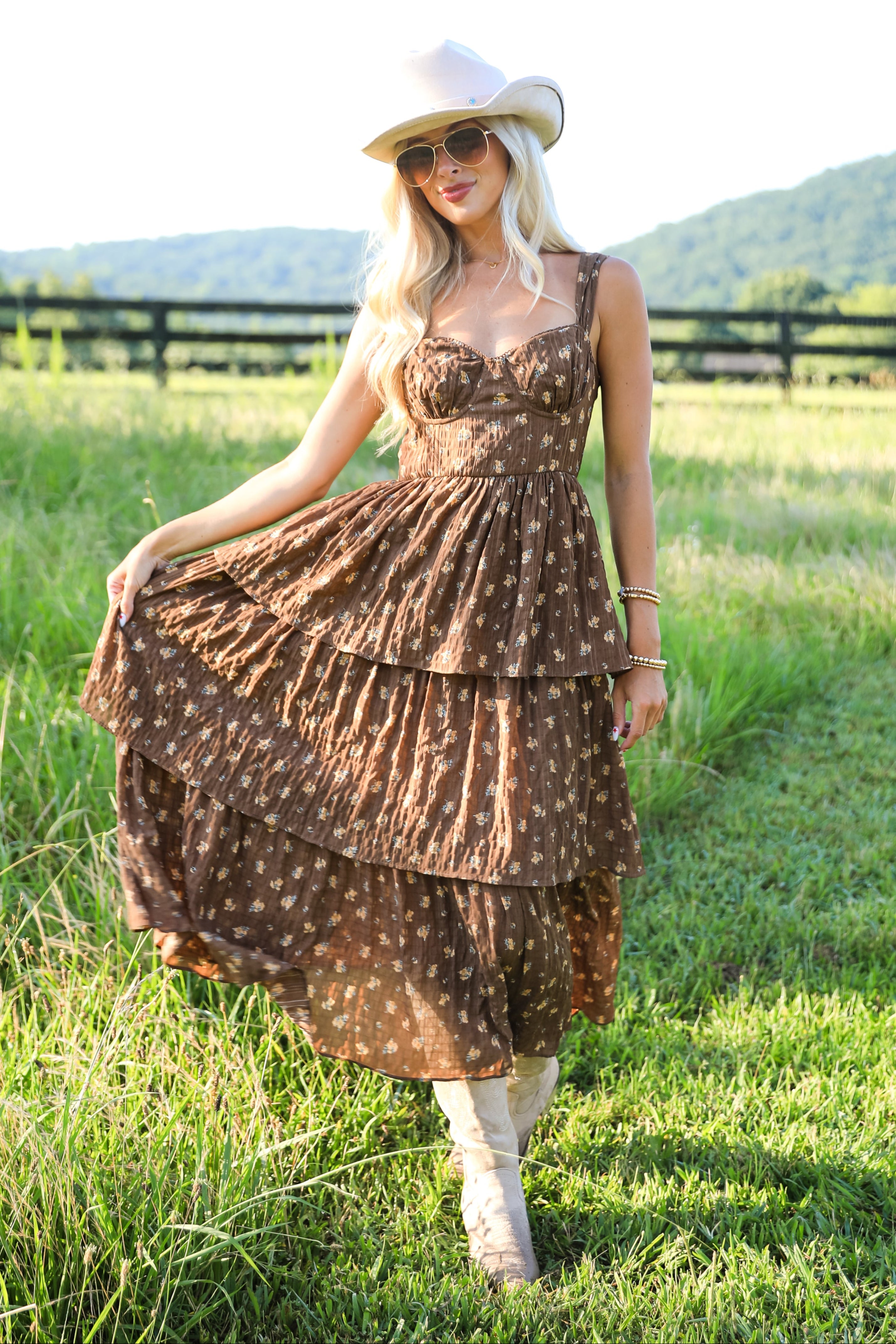 Woman in a brown tiered dress and white hat standing in a grassy field with mountains in the background