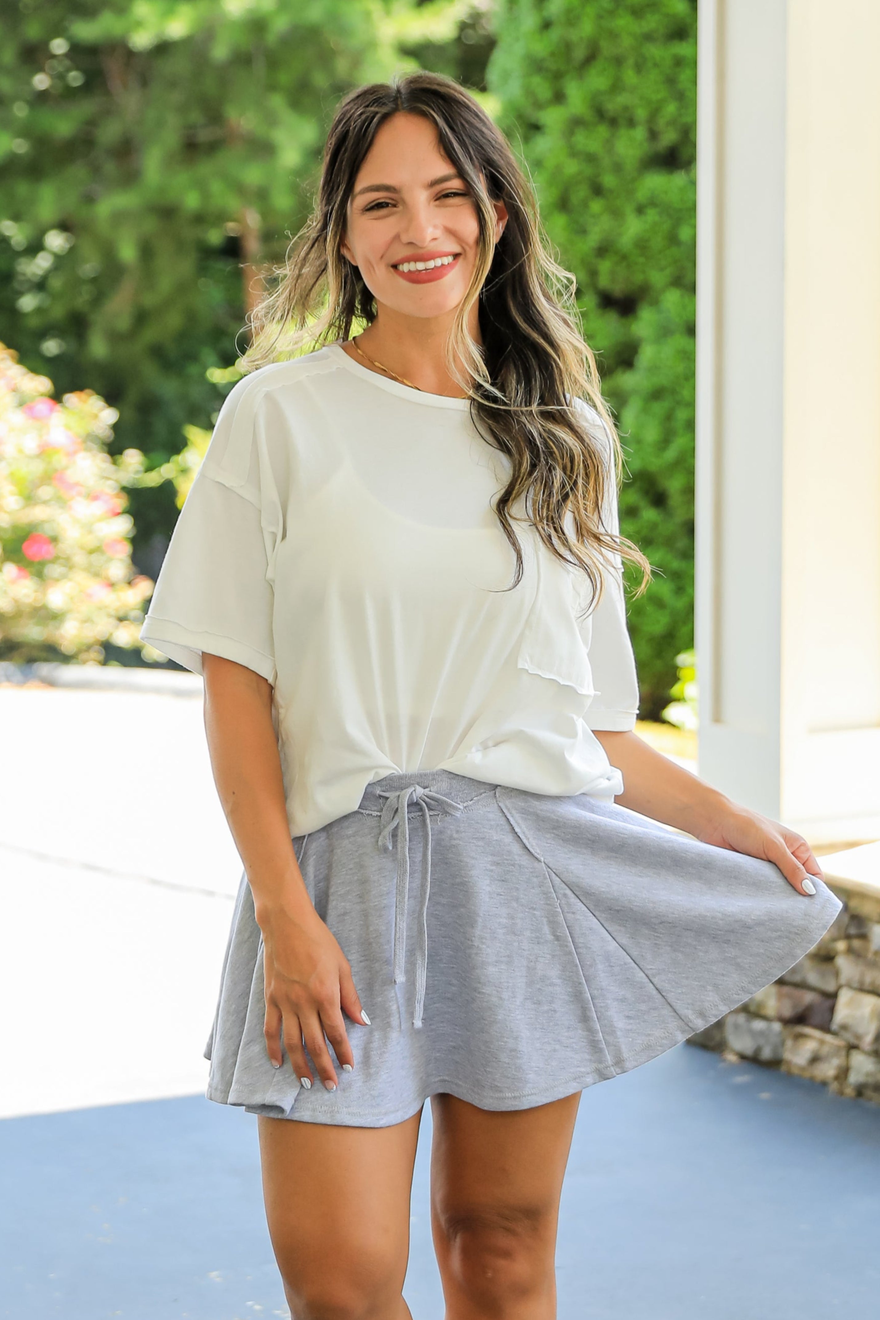 Woman in a white top and gray skirt standing on a patio with greenery in the background