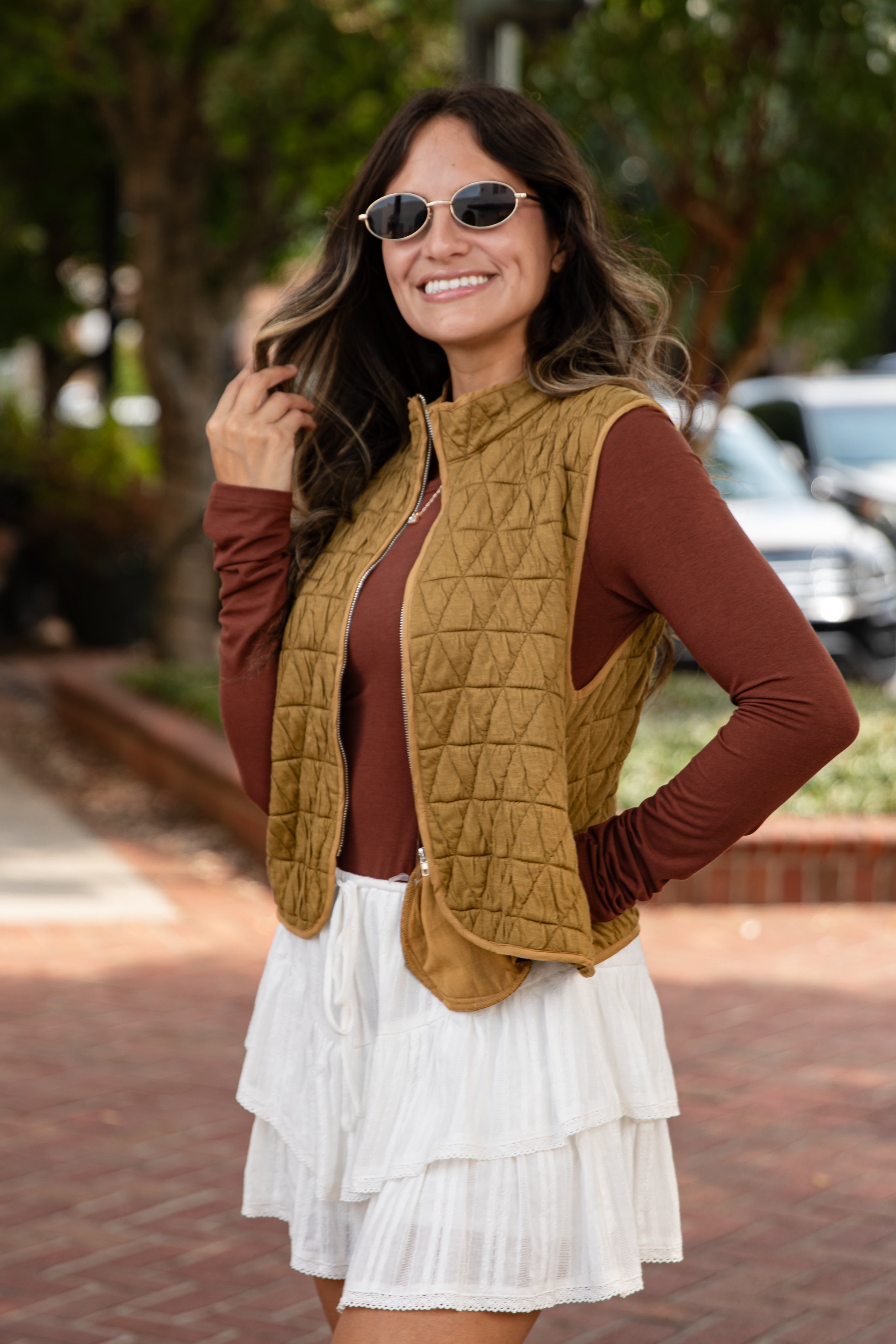 Woman wearing a mustard quilted vest, brown long-sleeve top, and white skirt outdoors.