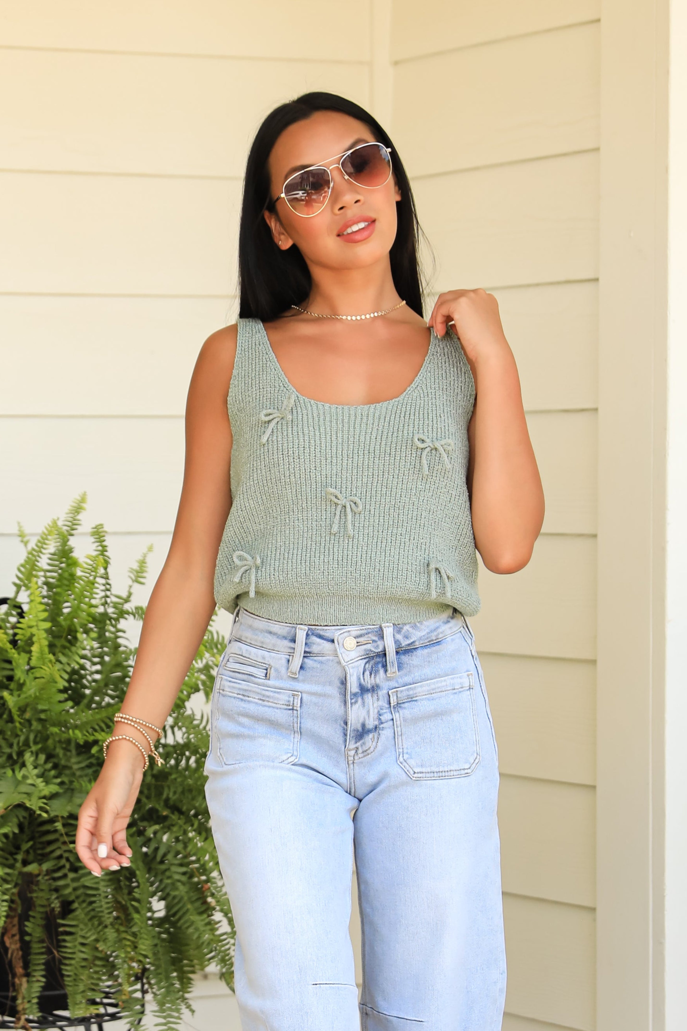 Woman in a green tank top and light blue jeans standing outdoors with plants and a lantern in the background.