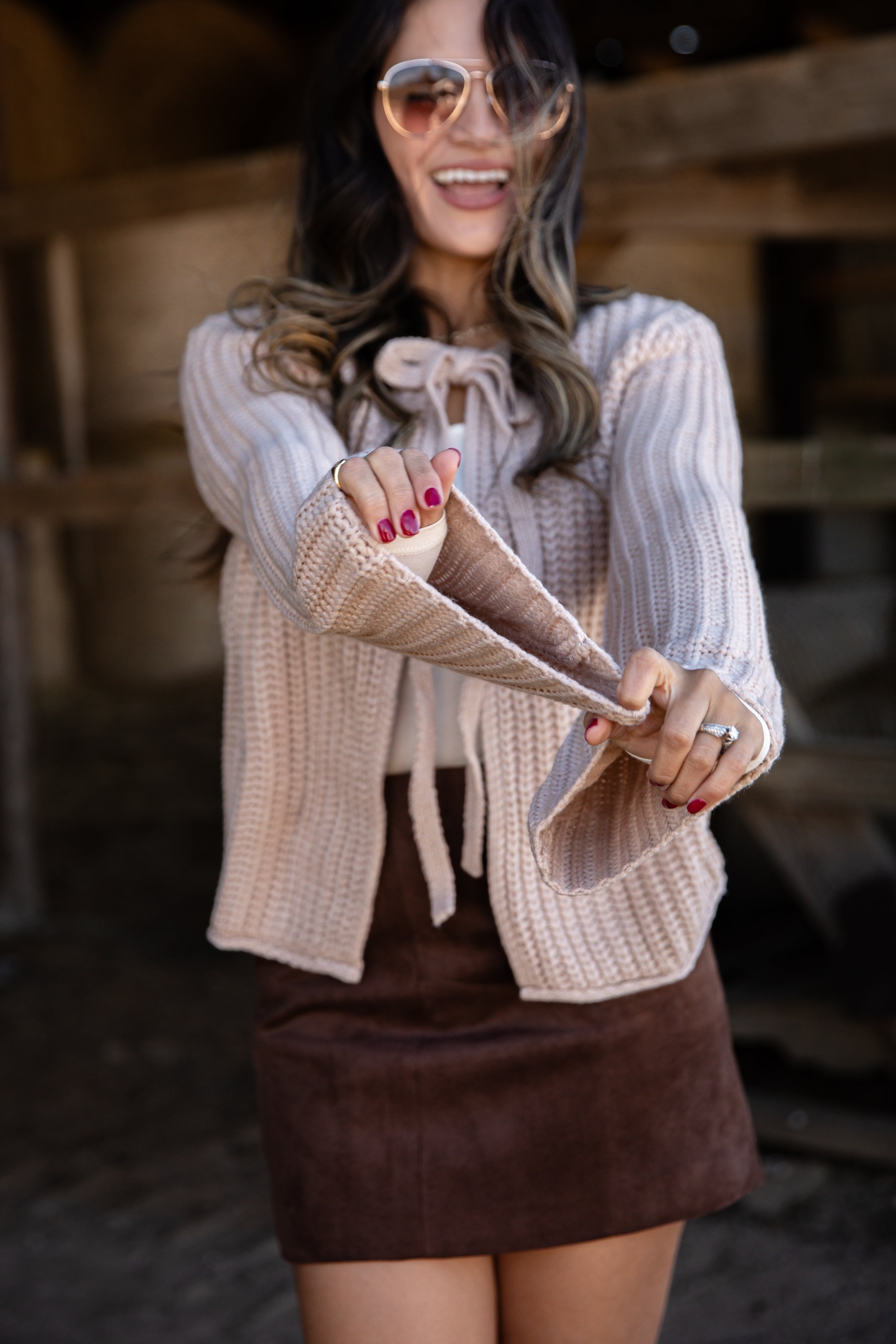 Woman wearing a beige cardigan over a white sweater with a brown skirt, standing in a rustic setting.