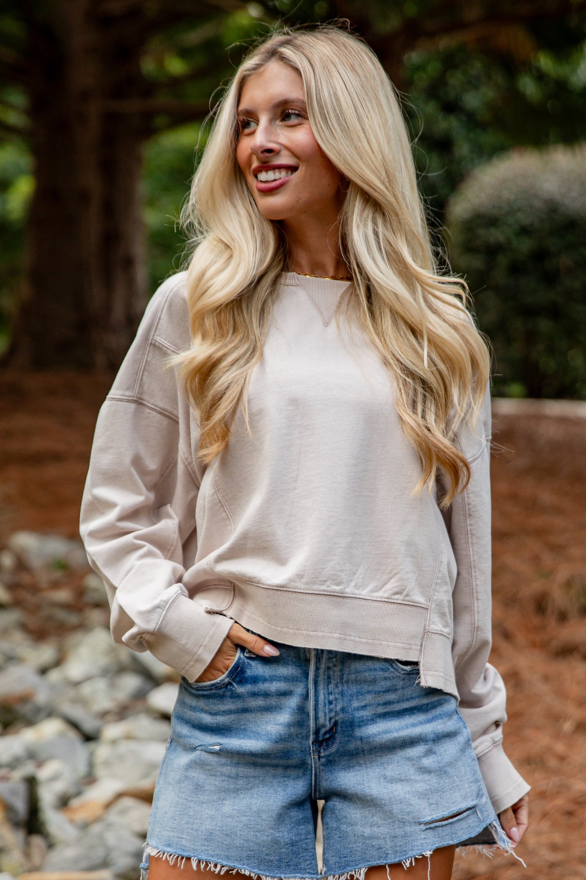 Woman in a light-colored sweatshirt and denim shorts standing outdoors with trees in the background