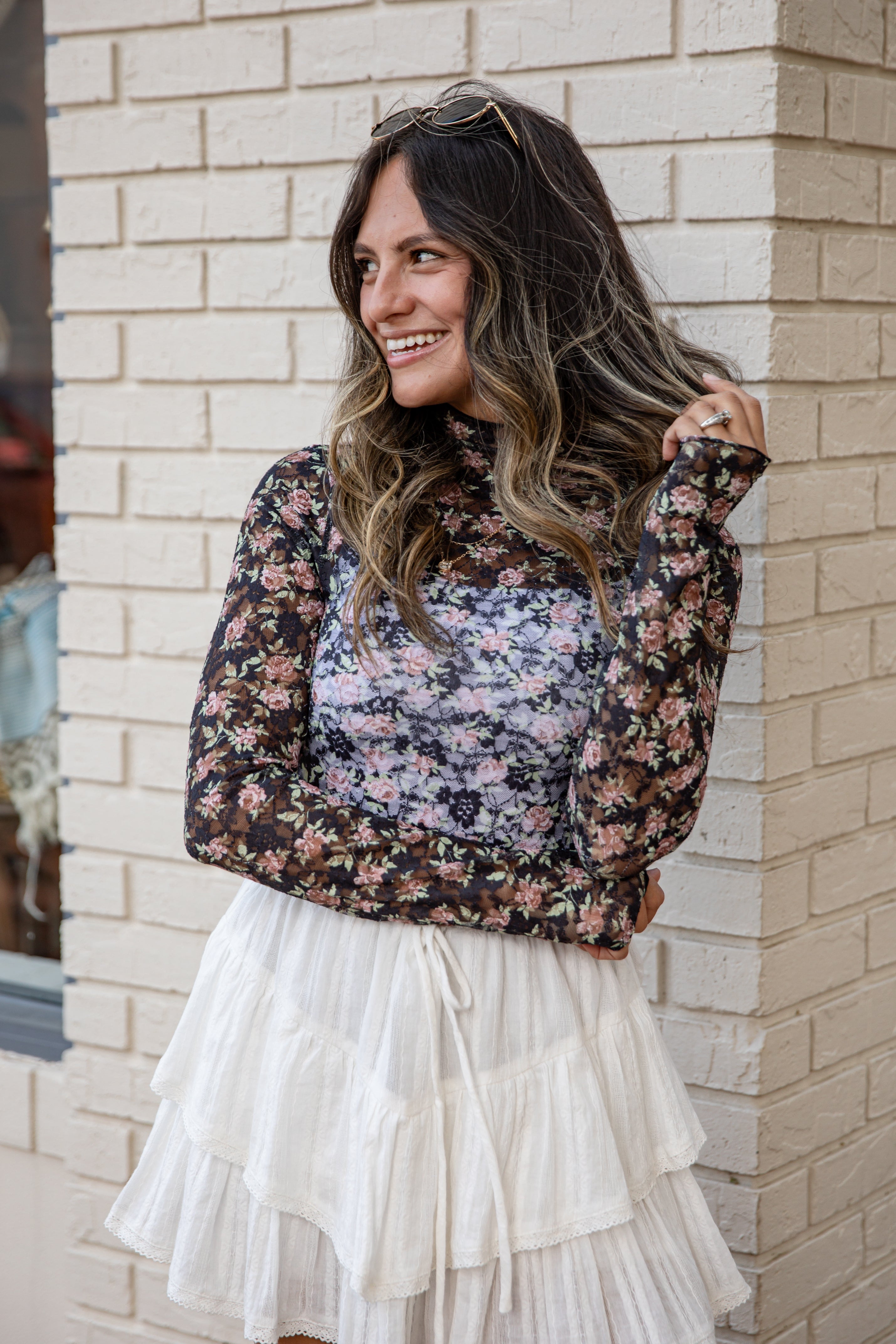 Woman wearing a floral blouse and white skirt against a brick wall.