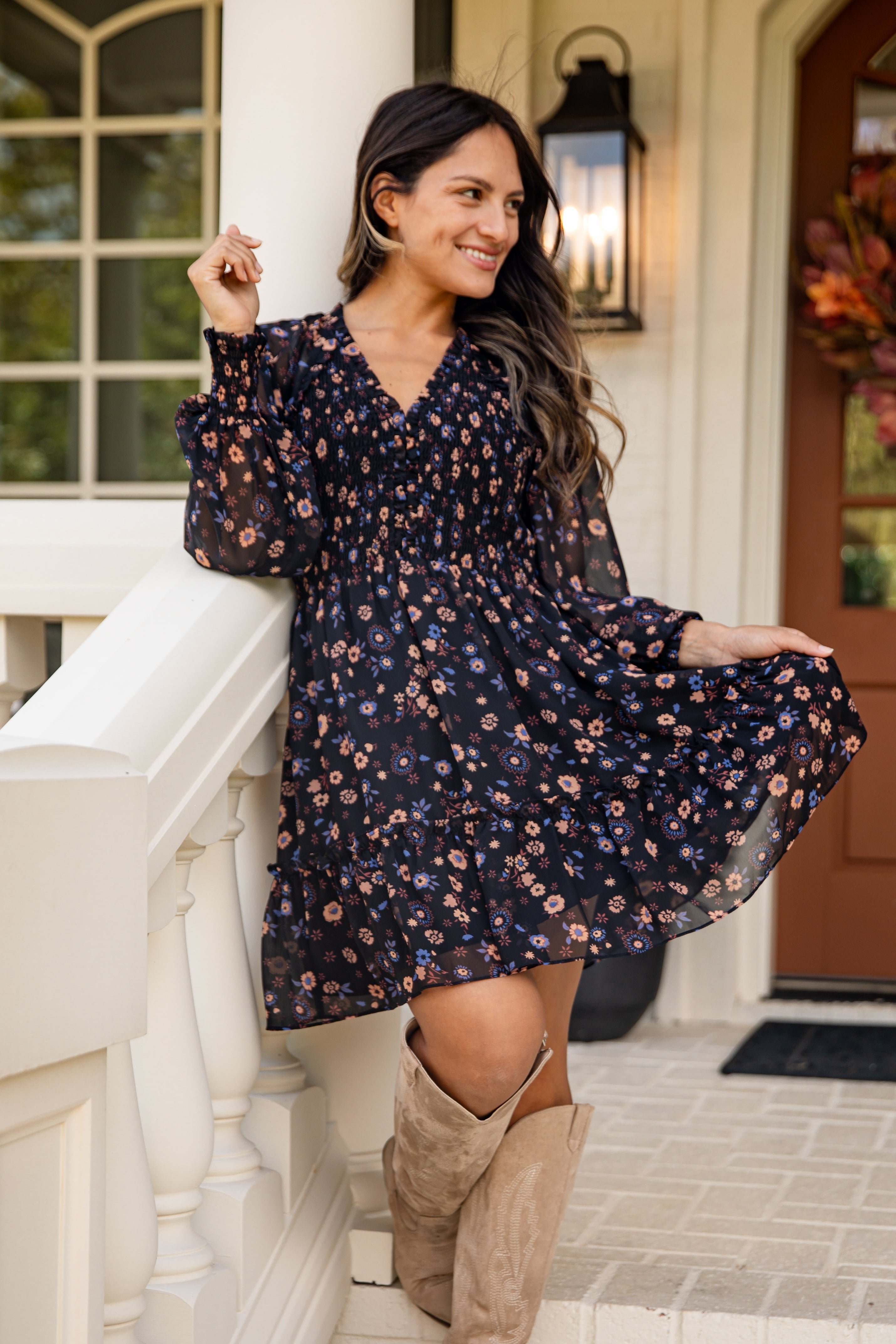 Woman in a floral dress and knee-high boots standing on a porch.
