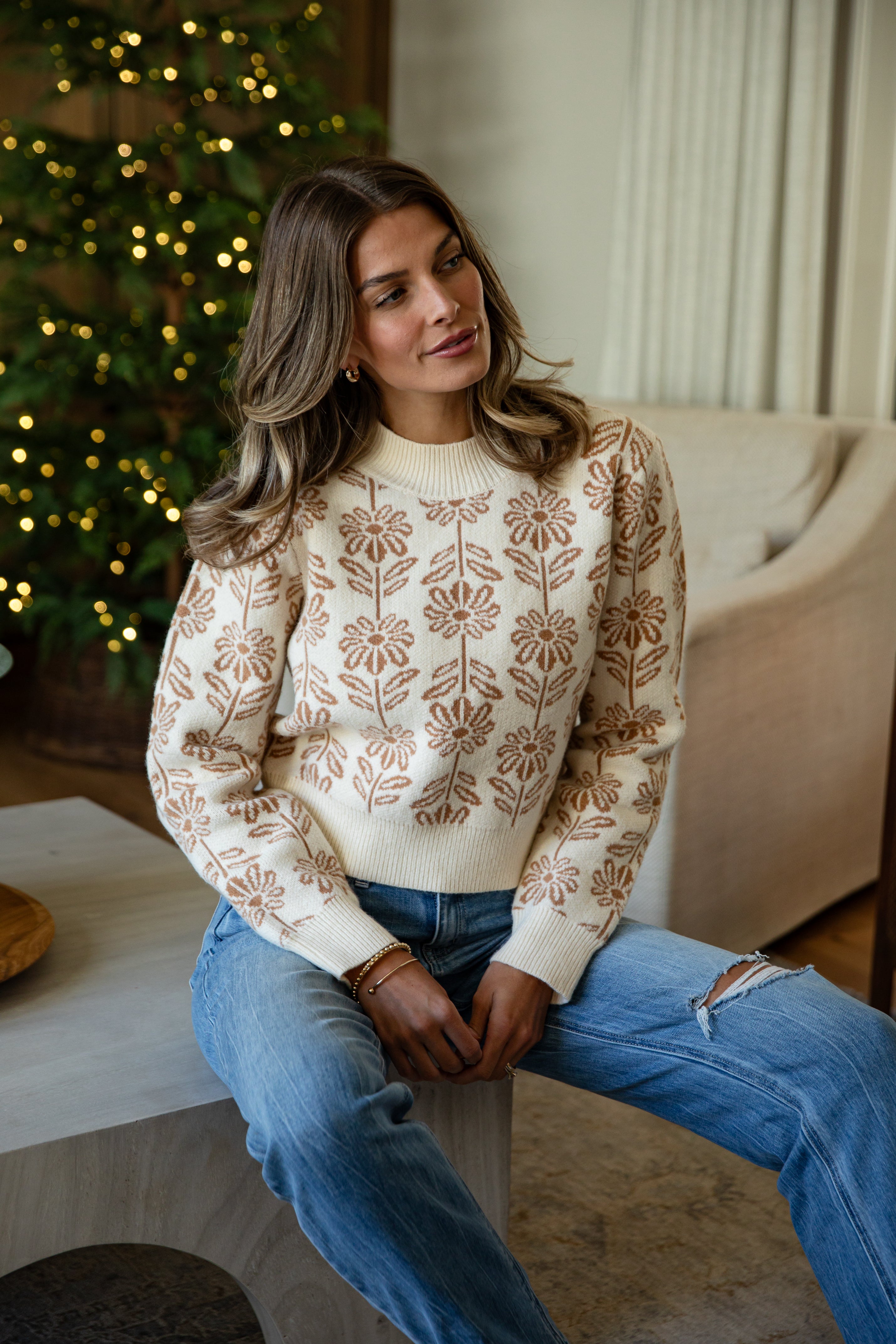 Woman wearing a patterned sweater and jeans sitting in a cozy living room with a Christmas tree.