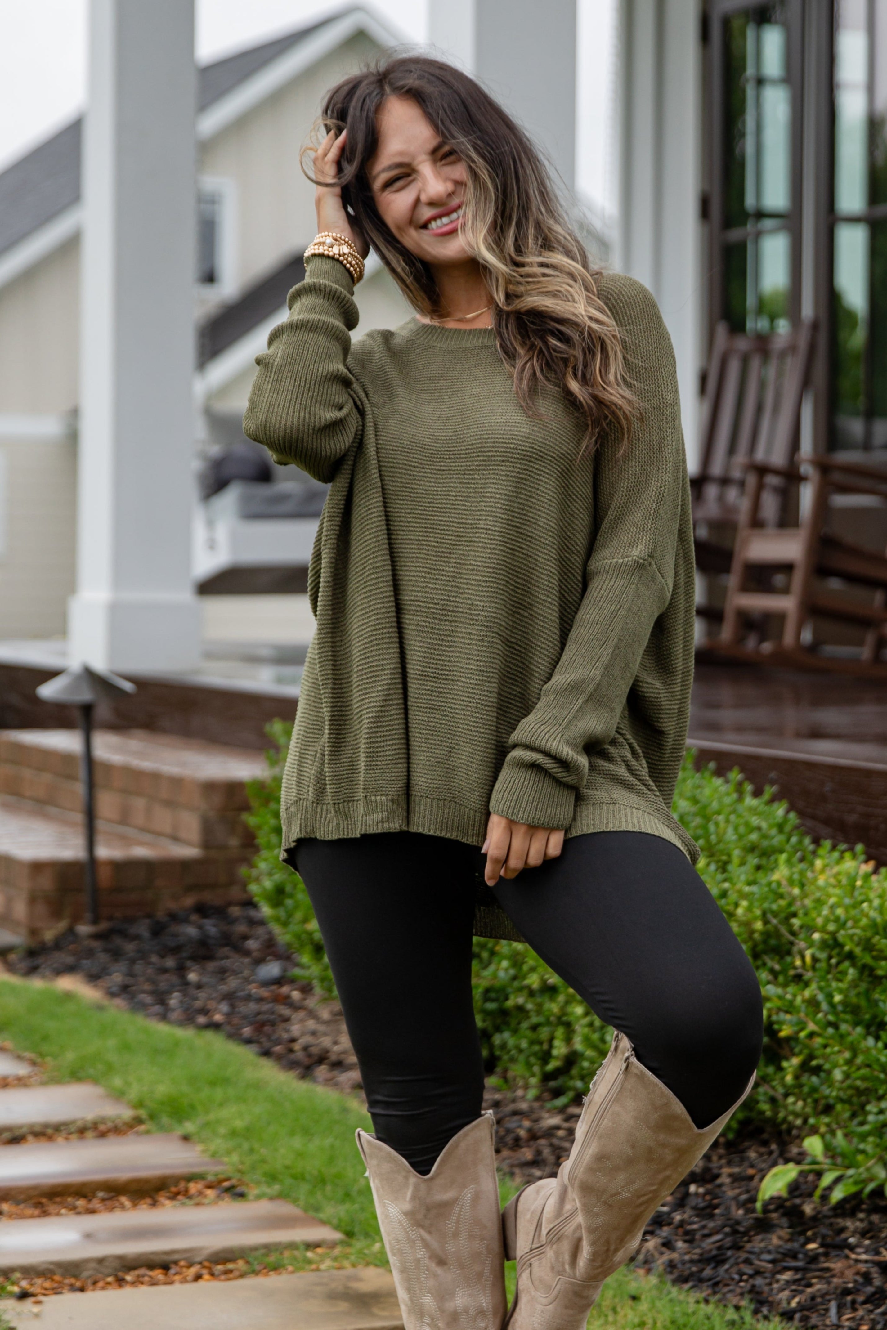 Woman in olive green sweater and beige knee-high boots walking on a stone path in front of a house.