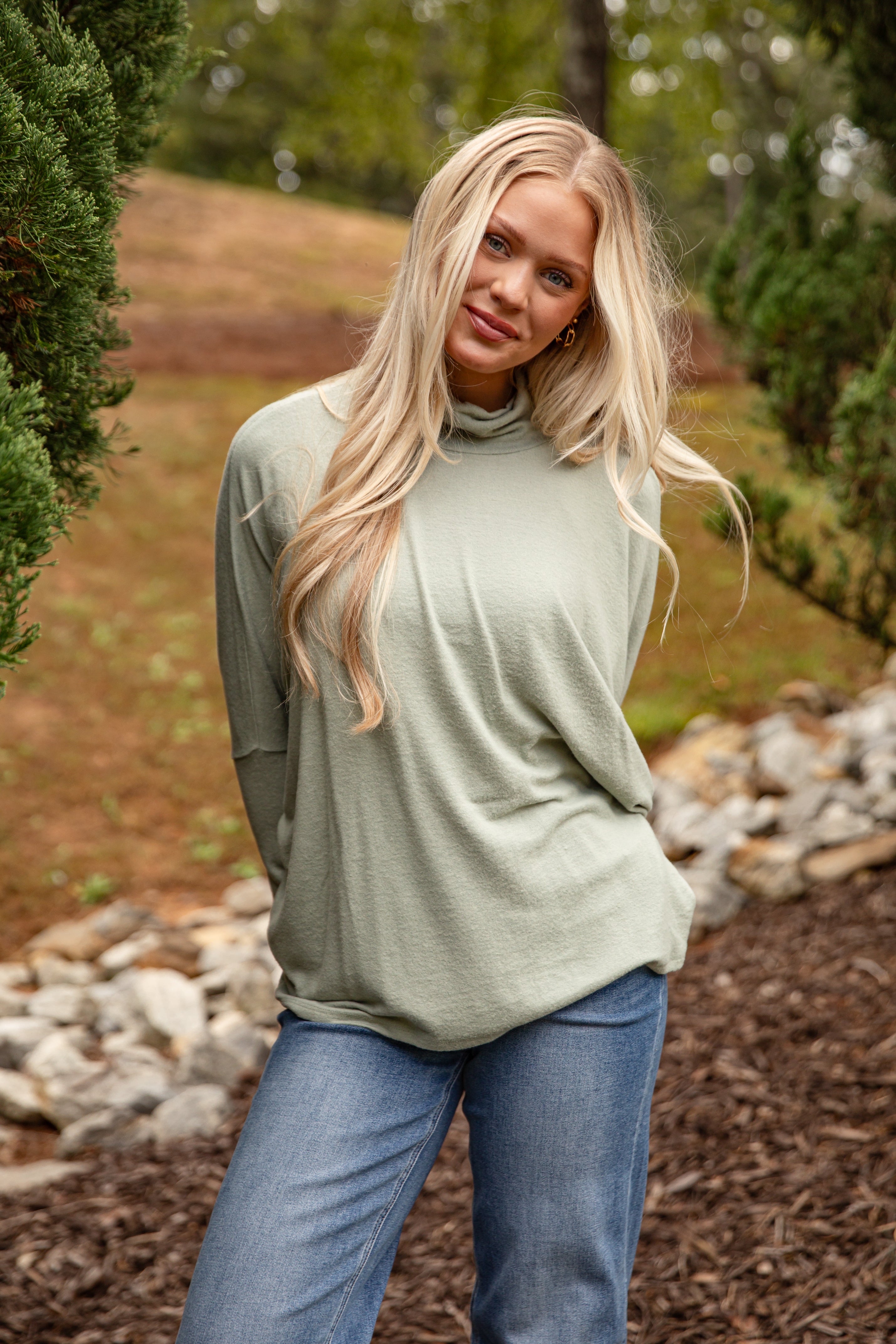 Woman wearing a green long-sleeve top and blue jeans standing outdoors with trees and rocks in the background.