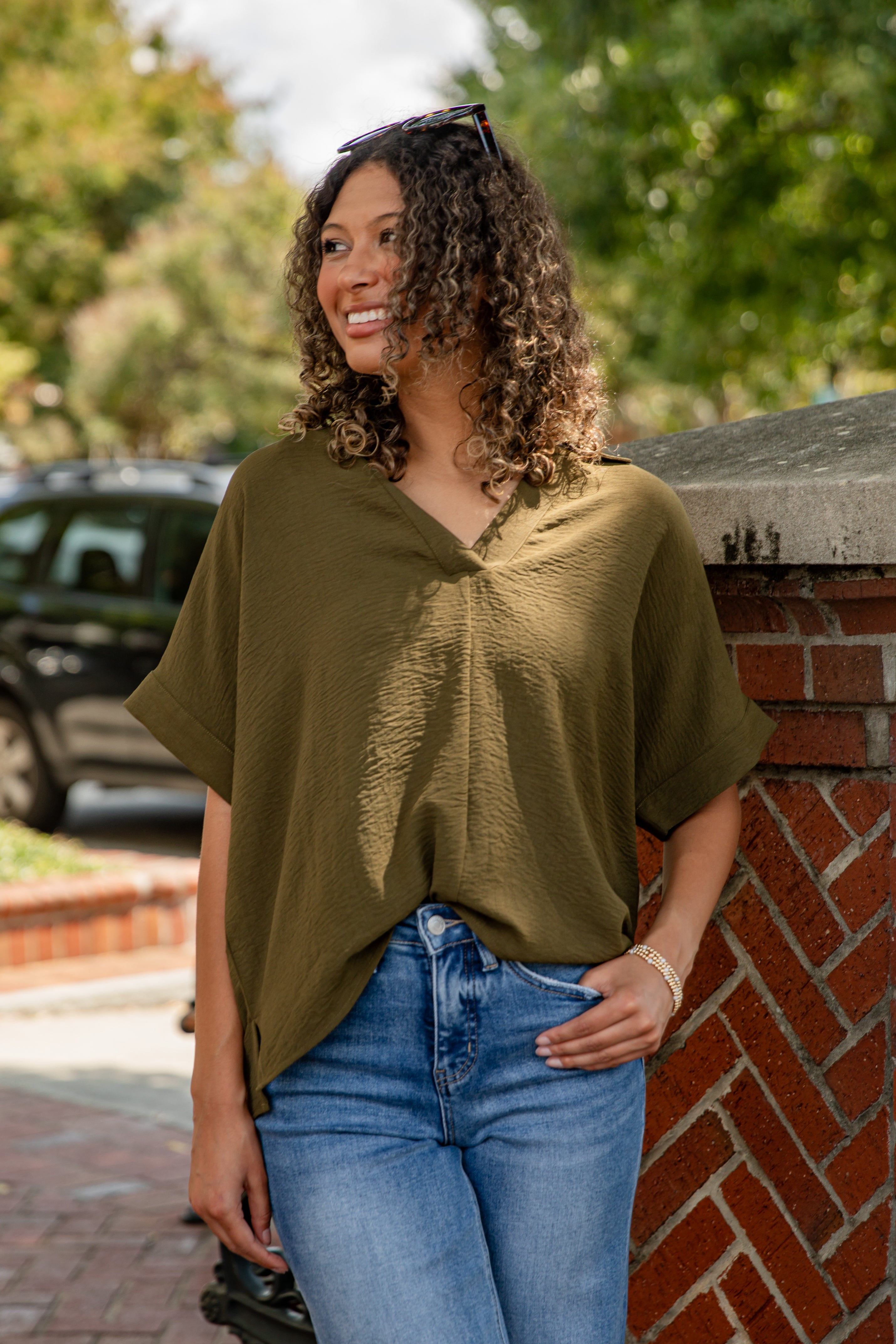Woman wearing an olive green top and blue jeans standing against a brick wall with trees in the background.