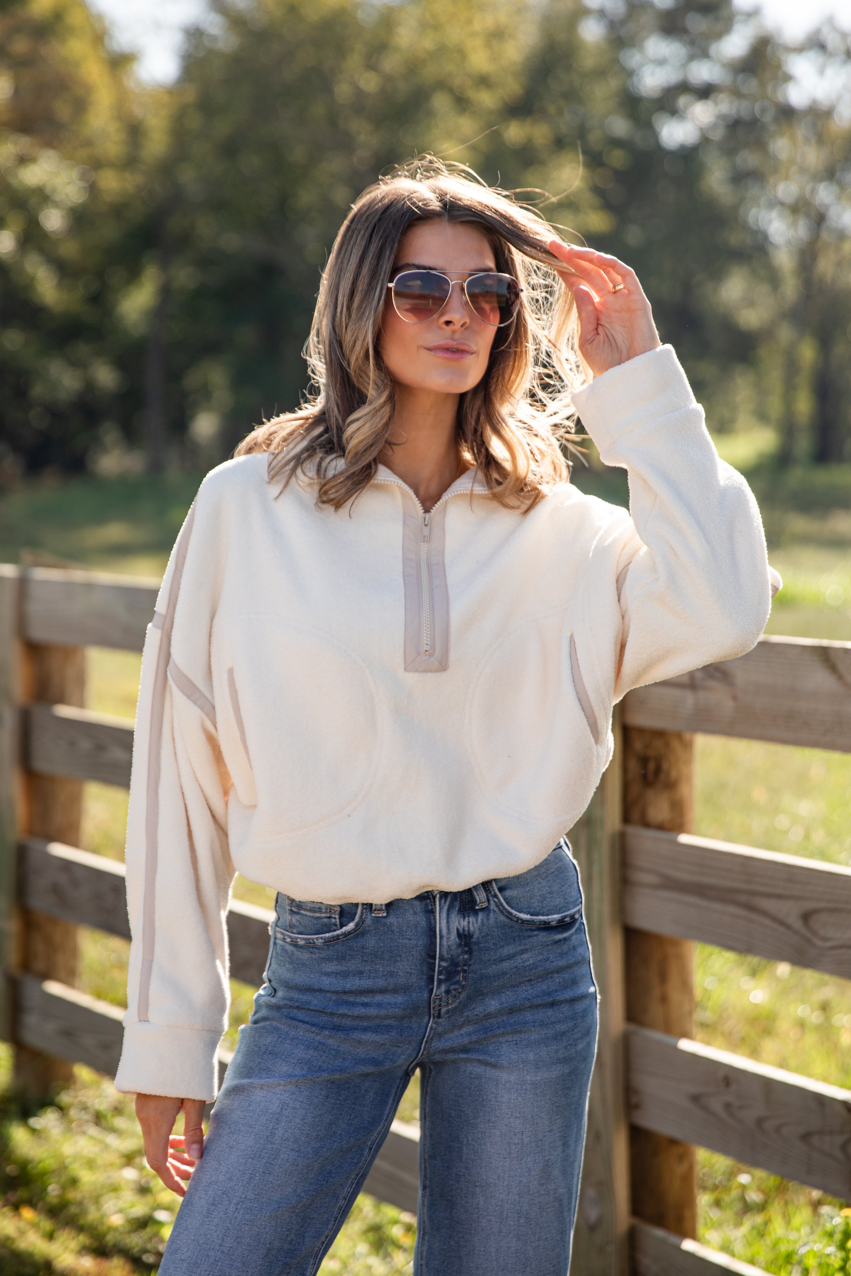 Woman wearing a cream blouse and blue jeans standing in front of a wooden fence with trees in the background.