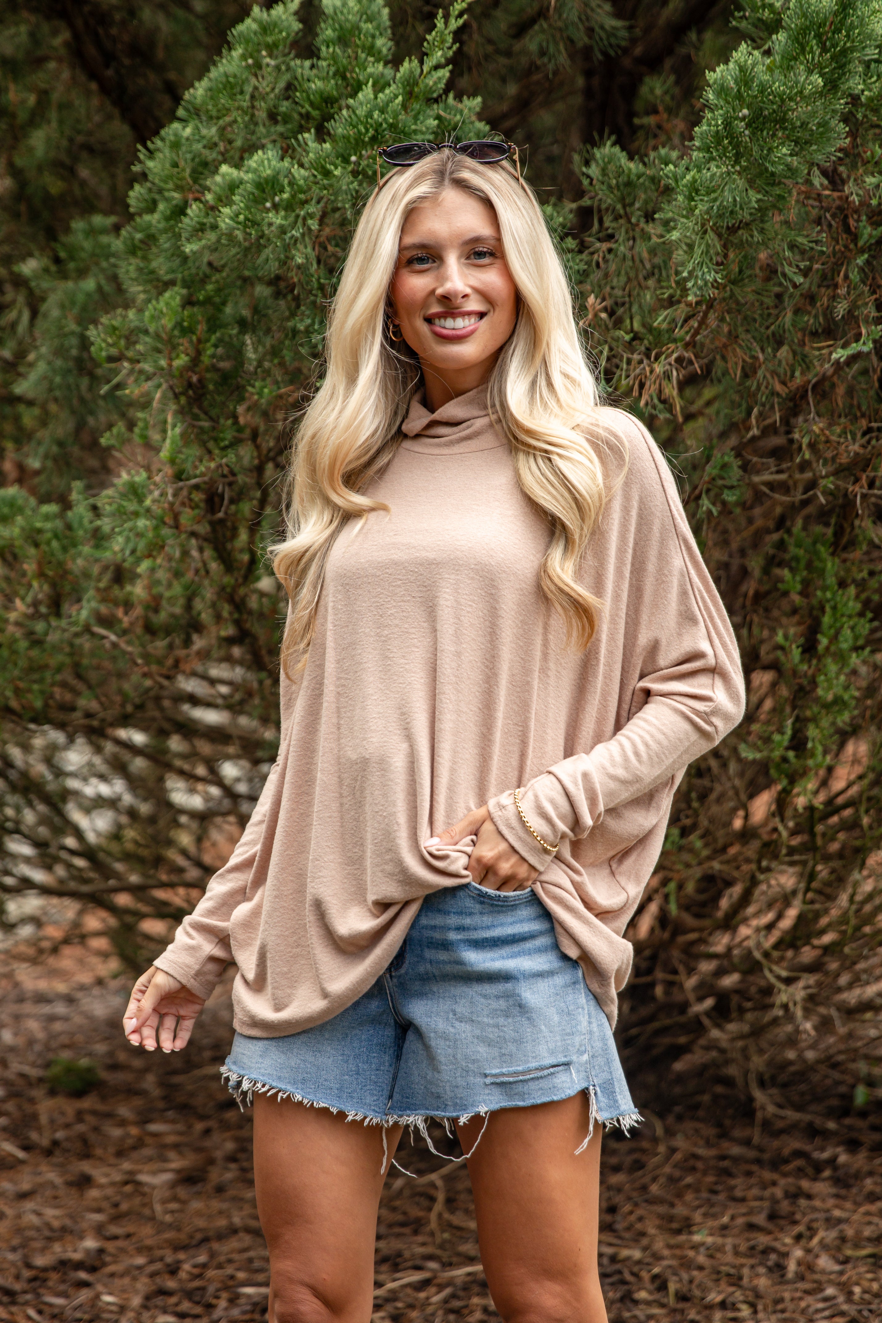 Woman wearing a beige long-sleeve top and denim shorts standing outdoors with greenery in the background