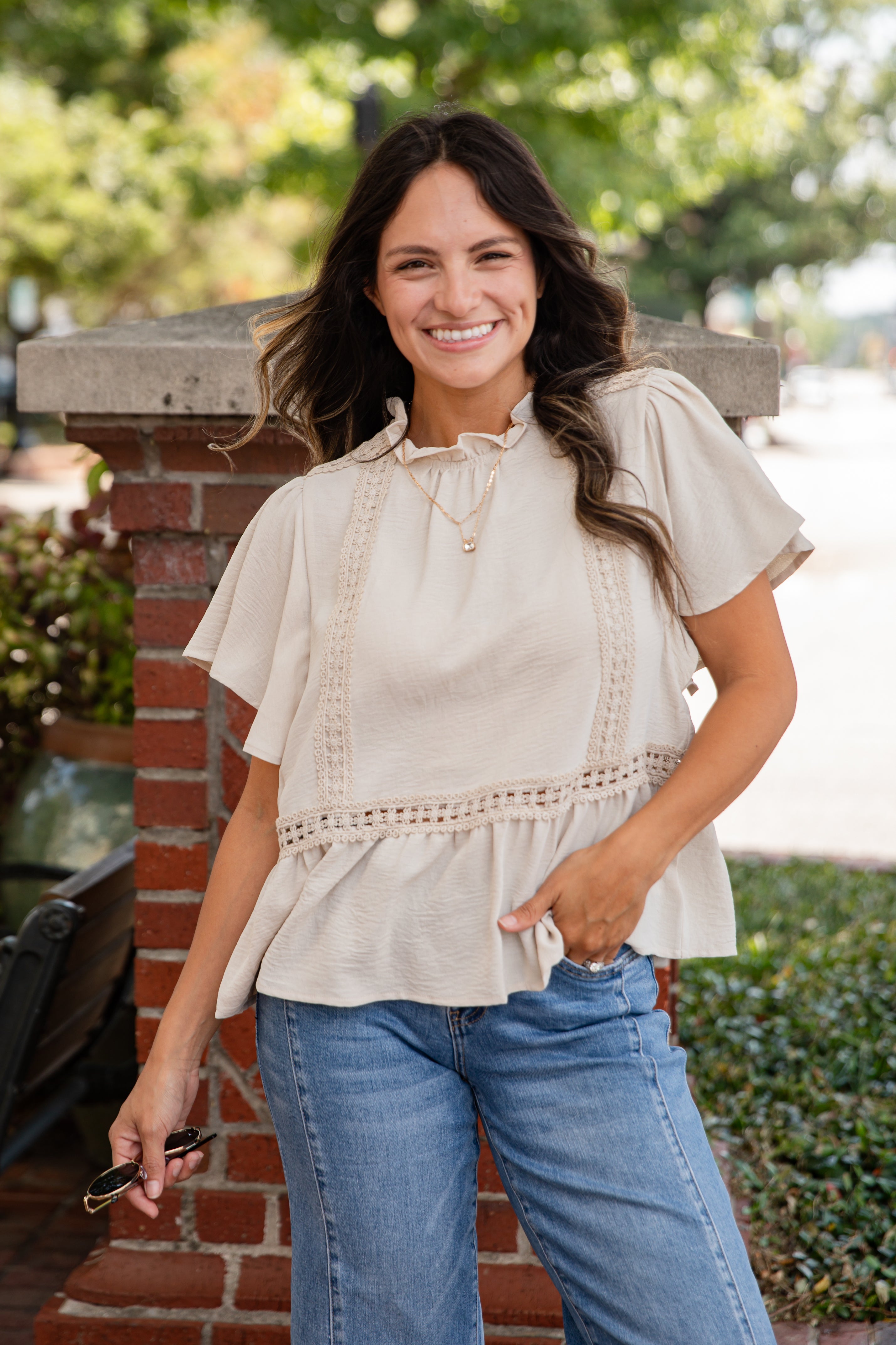 Woman wearing a beige blouse and blue jeans standing outdoors with greenery in the background