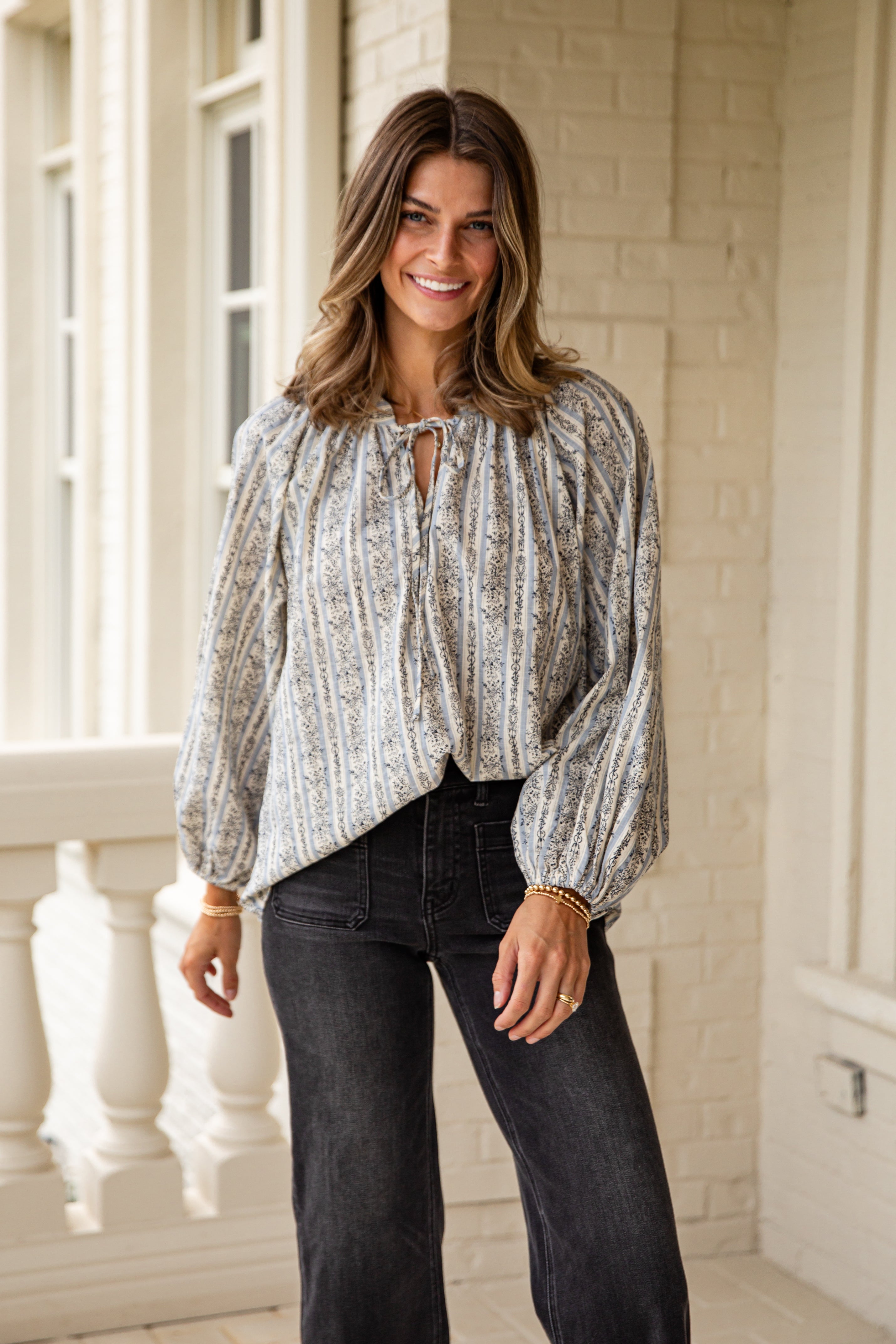 Woman wearing a patterned blouse and dark jeans standing in a bright room with large windows.