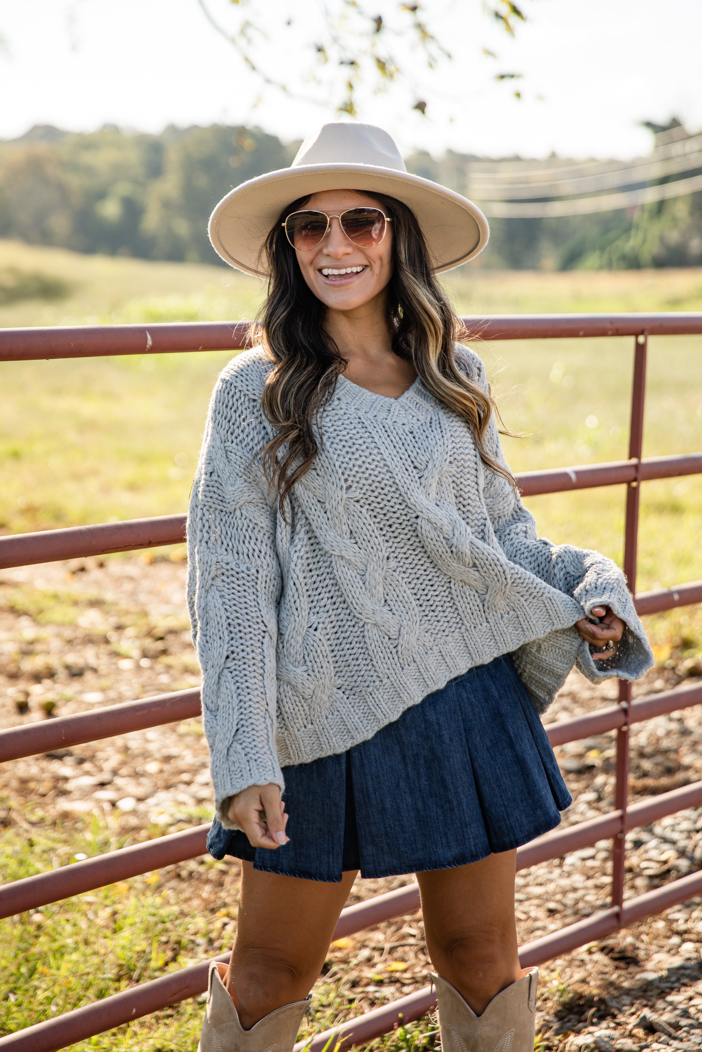 Woman wearing a light gray cable knit sweater, denim shorts, beige cowboy boots, and a wide-brimmed hat outdoors.