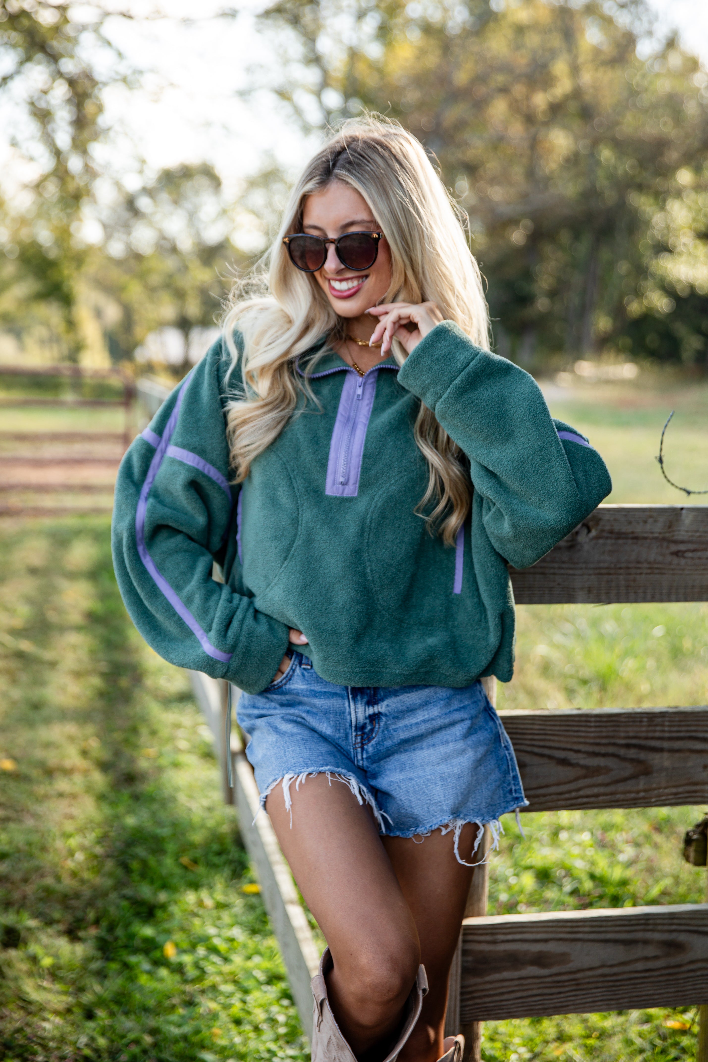 Woman wearing a green sweater with purple accents and denim shorts standing outdoors.