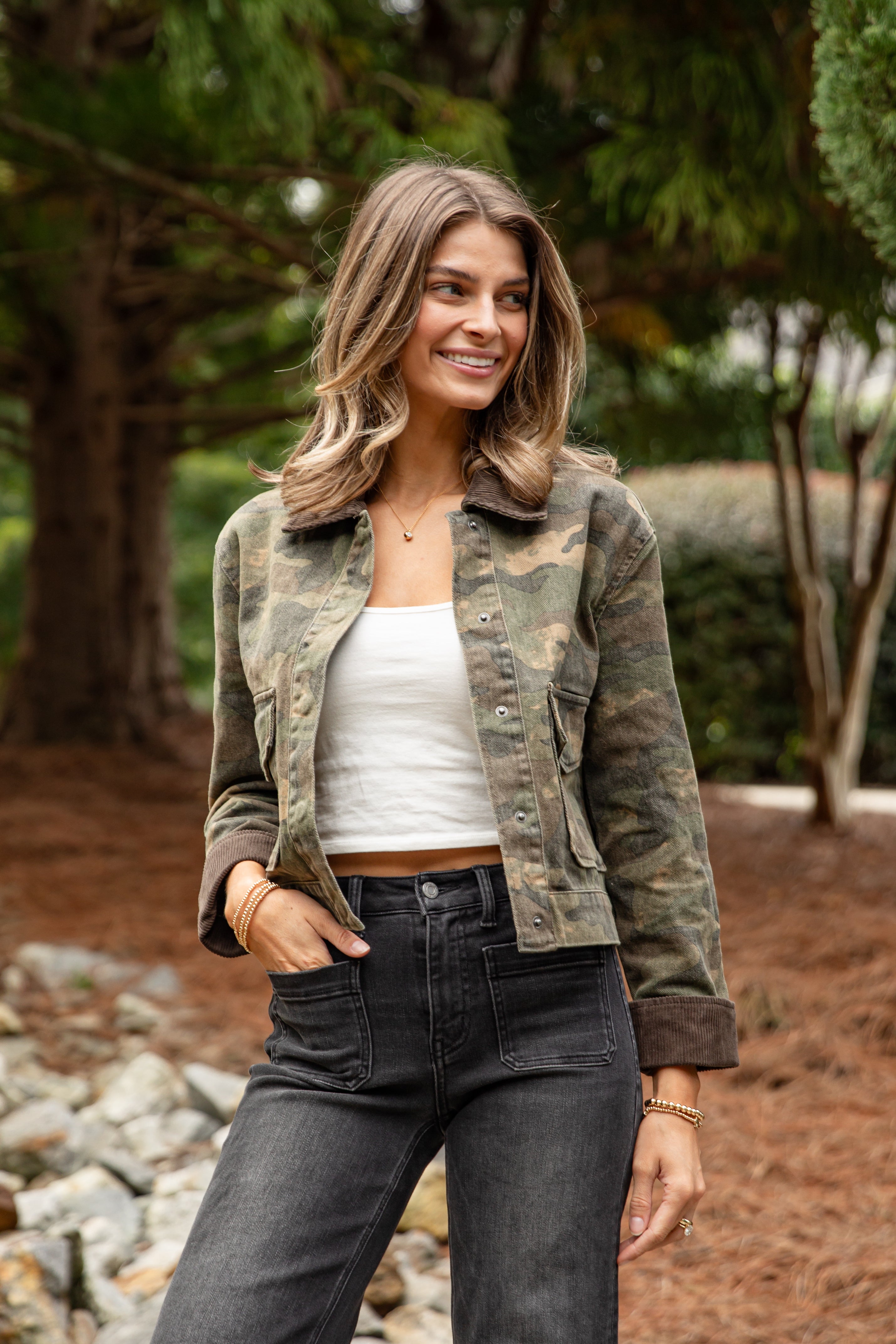 Woman wearing a camouflage jacket, white top, and dark jeans outdoors.