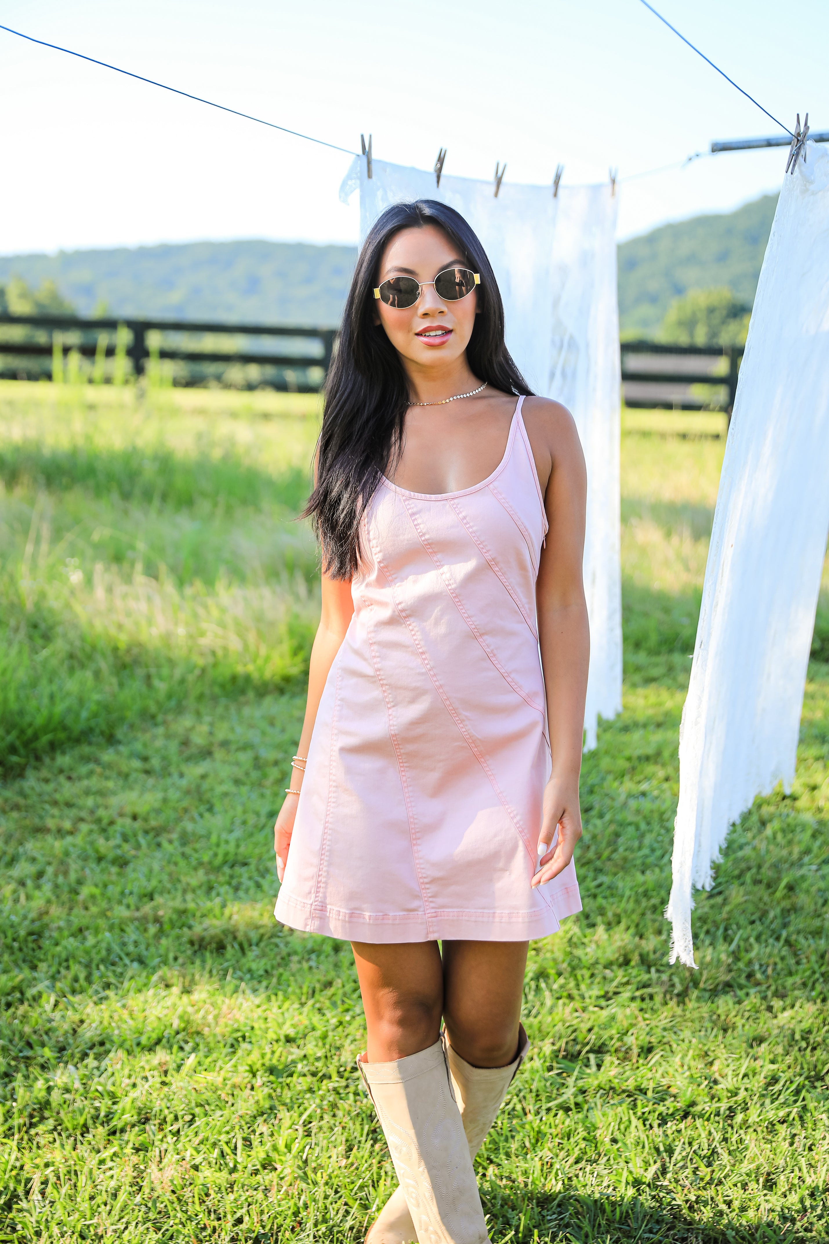 Woman in a pink dress and beige boots standing in a grassy field with mountains in the background.