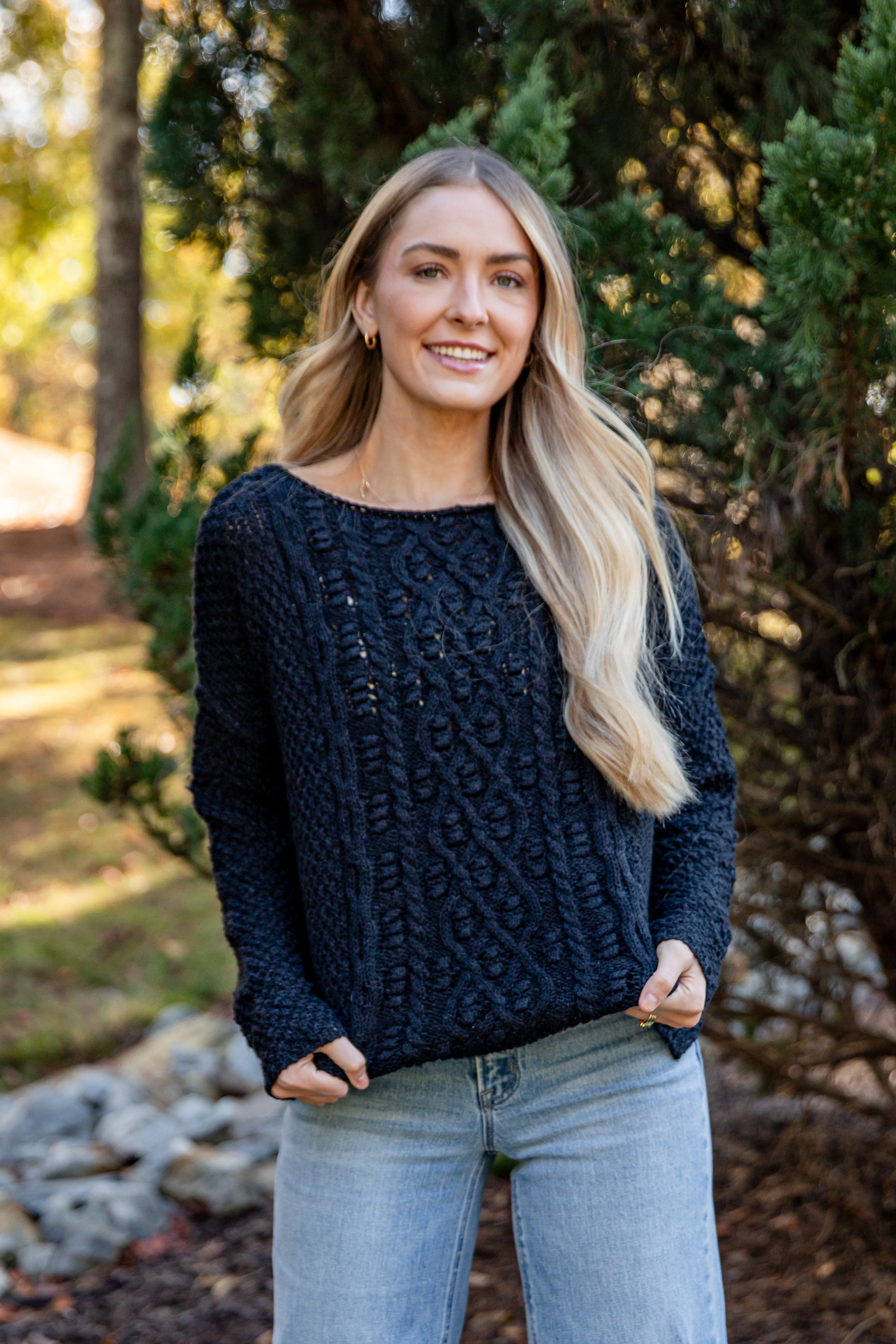 Woman wearing a dark blue sweater and light blue jeans standing outdoors with trees in the background