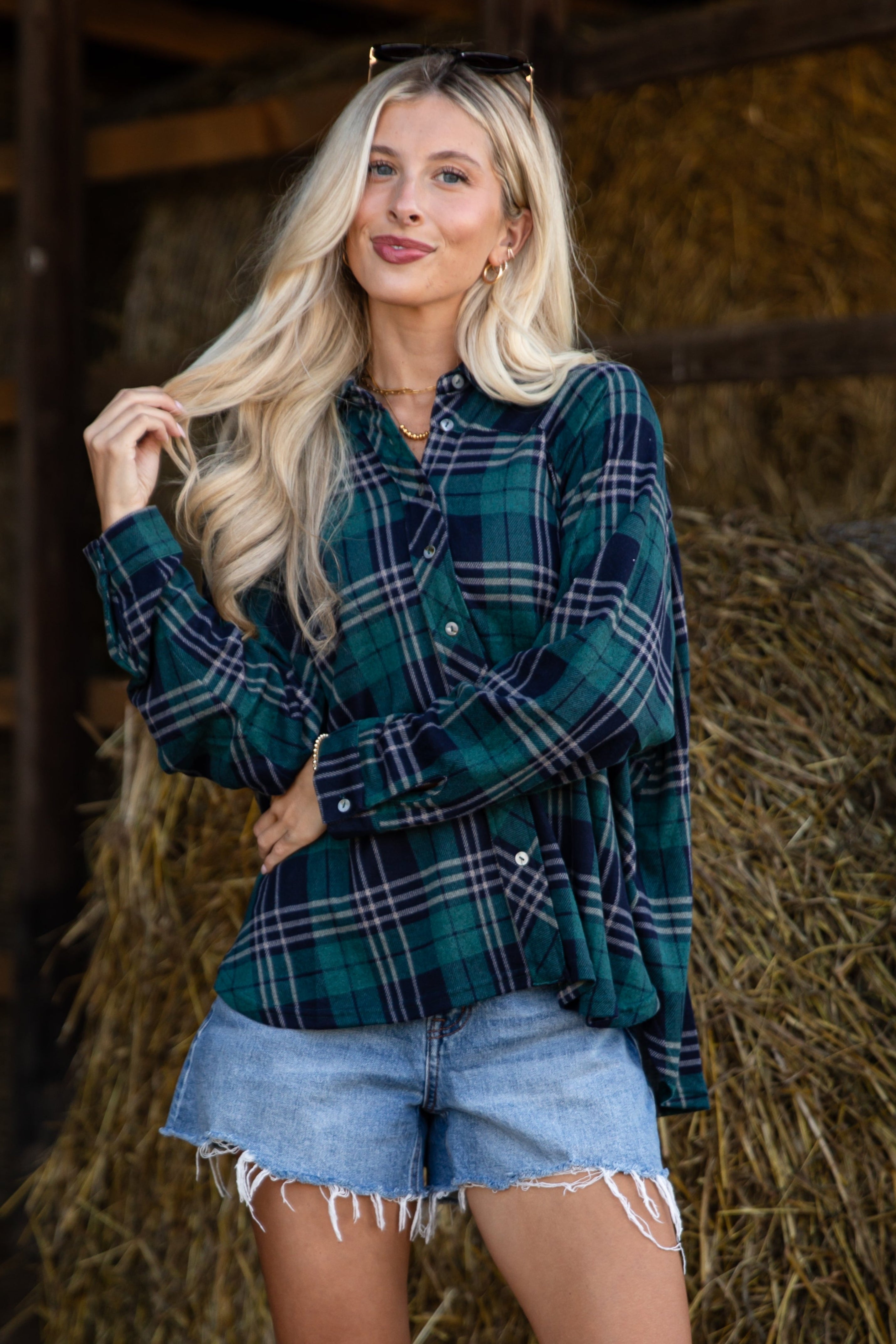 Woman wearing a plaid shirt and denim shorts standing in a barn with hay bales.