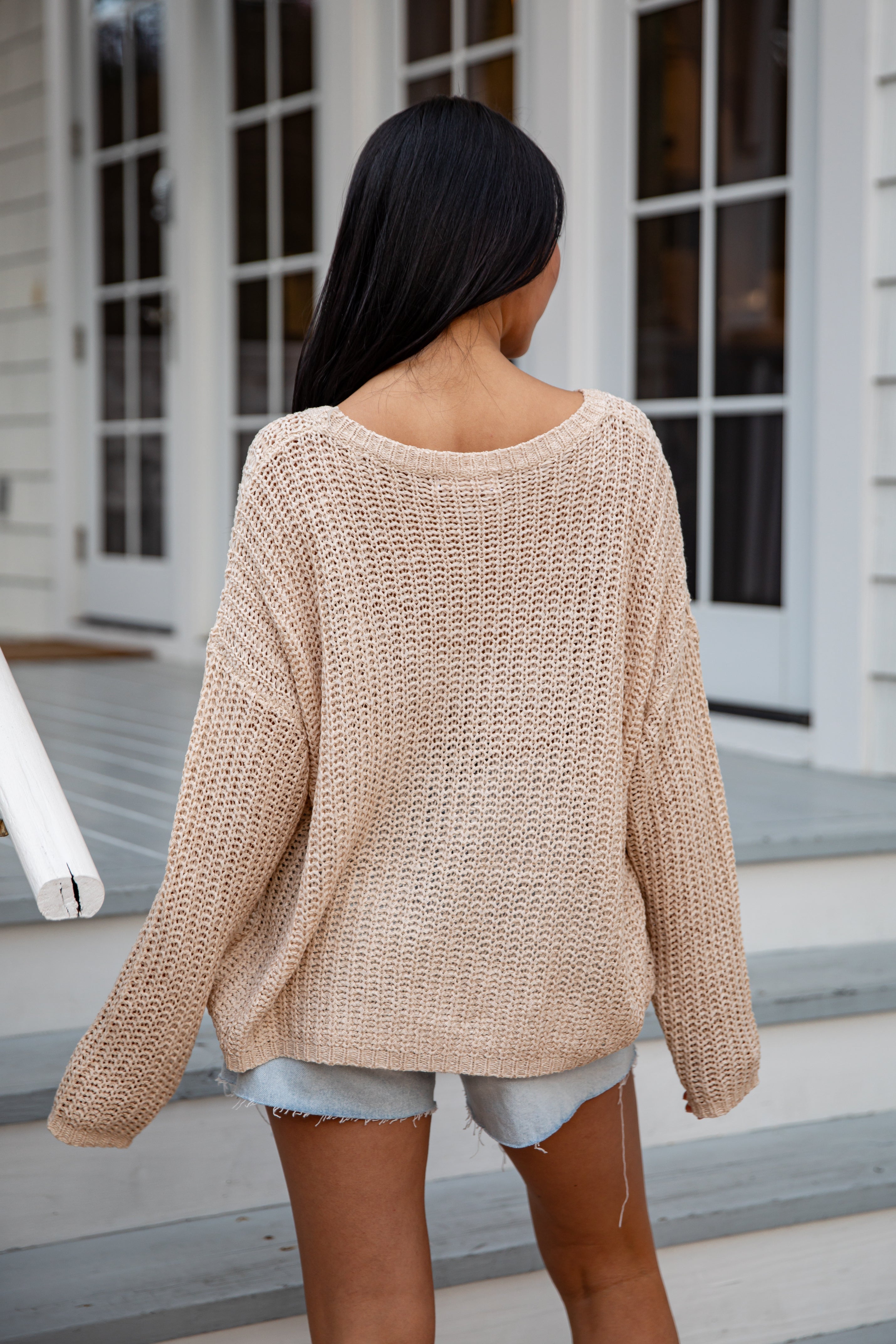 Woman wearing a beige knitted sweater and denim shorts standing on a porch.