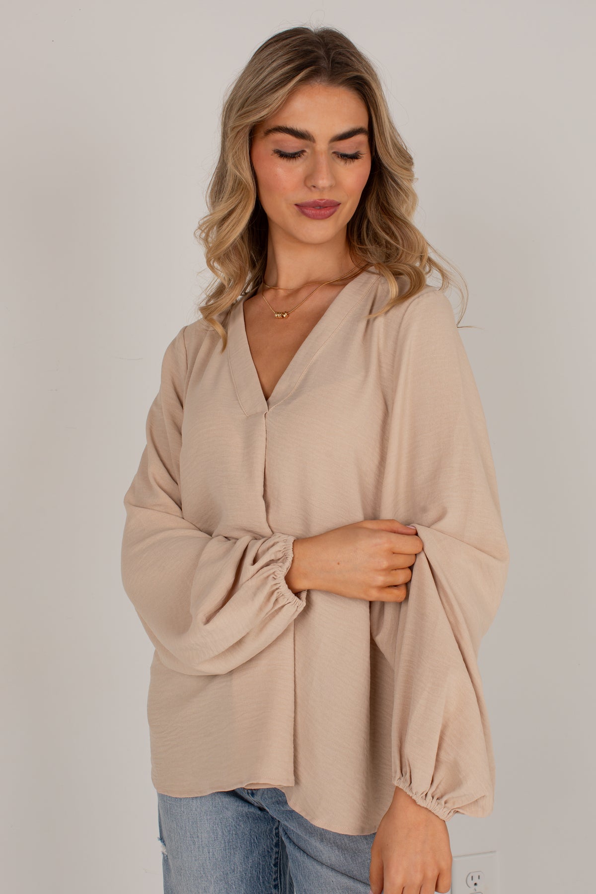 A woman wearing a beige long-sleeve blouse with balloon sleeves and a V-neckline, paired with blue jeans, photographed against a neutral light gray background.