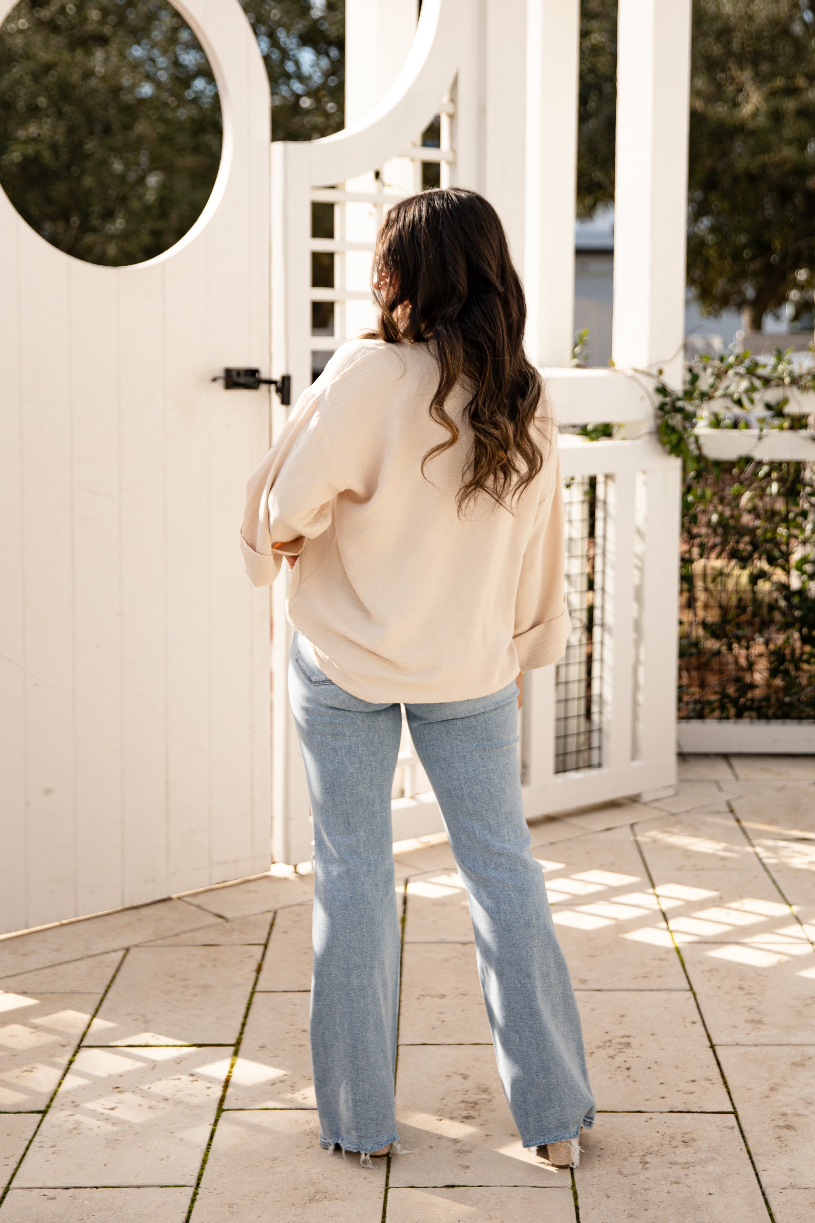 Woman in a beige sweater and light blue jeans standing on a patio with a white gate and greenery in the background.