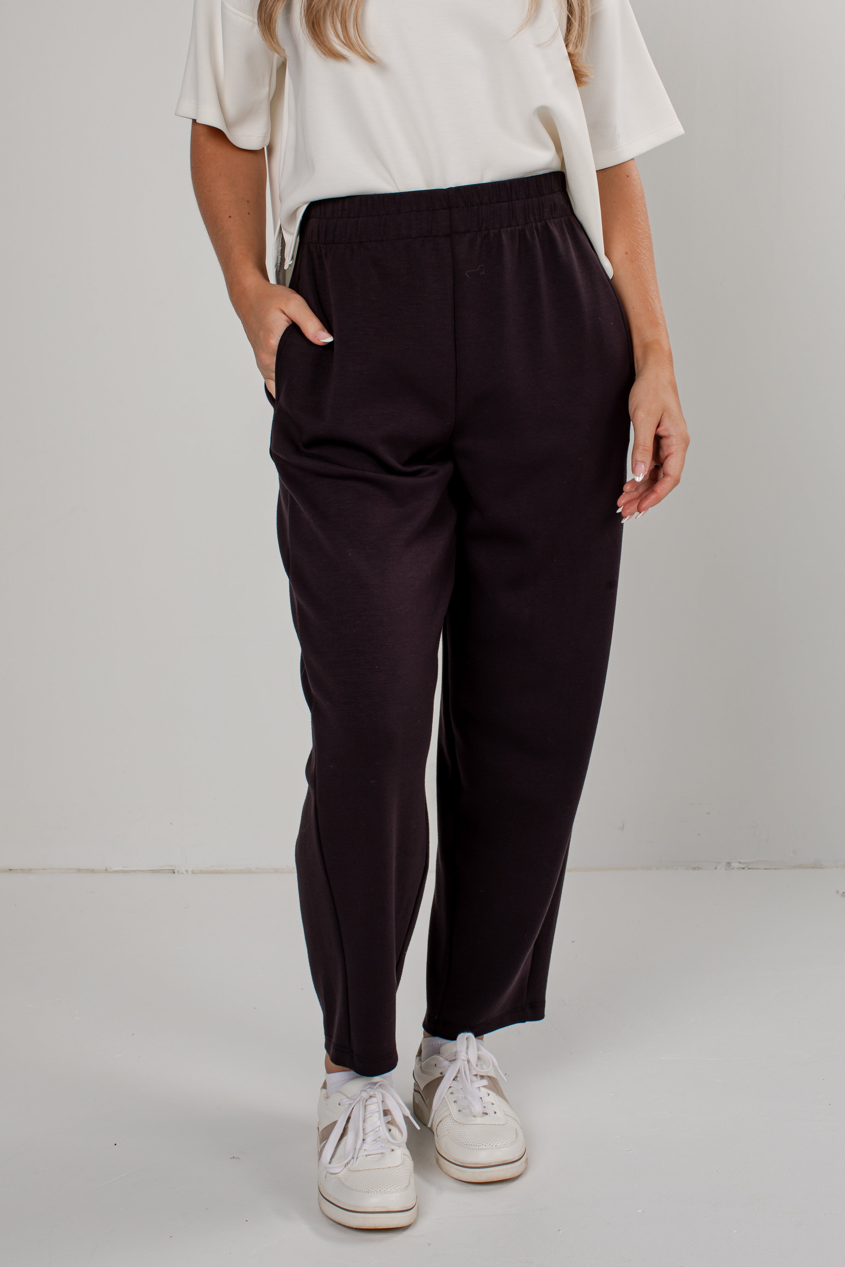 A person wearing dark black-colored wide-leg sweatpants with an elastic waistband and side pockets, styled with a white t-shirt and white sneakers against a plain light gray studio background.