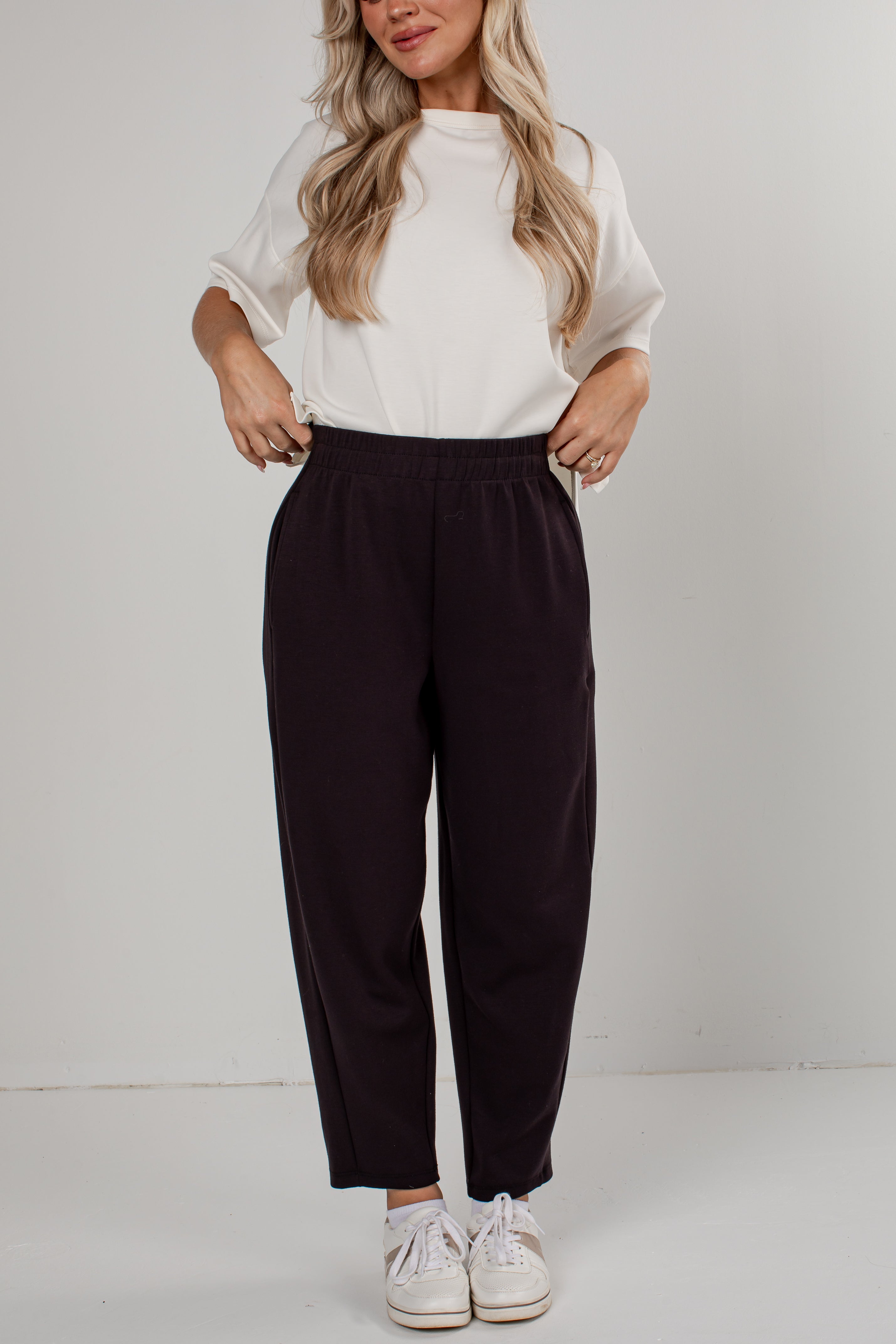 A woman modeling a pair of dark plum-colored wide-leg cropped pants, paired with a white short-sleeve top and white sneakers, against a plain light gray studio background.