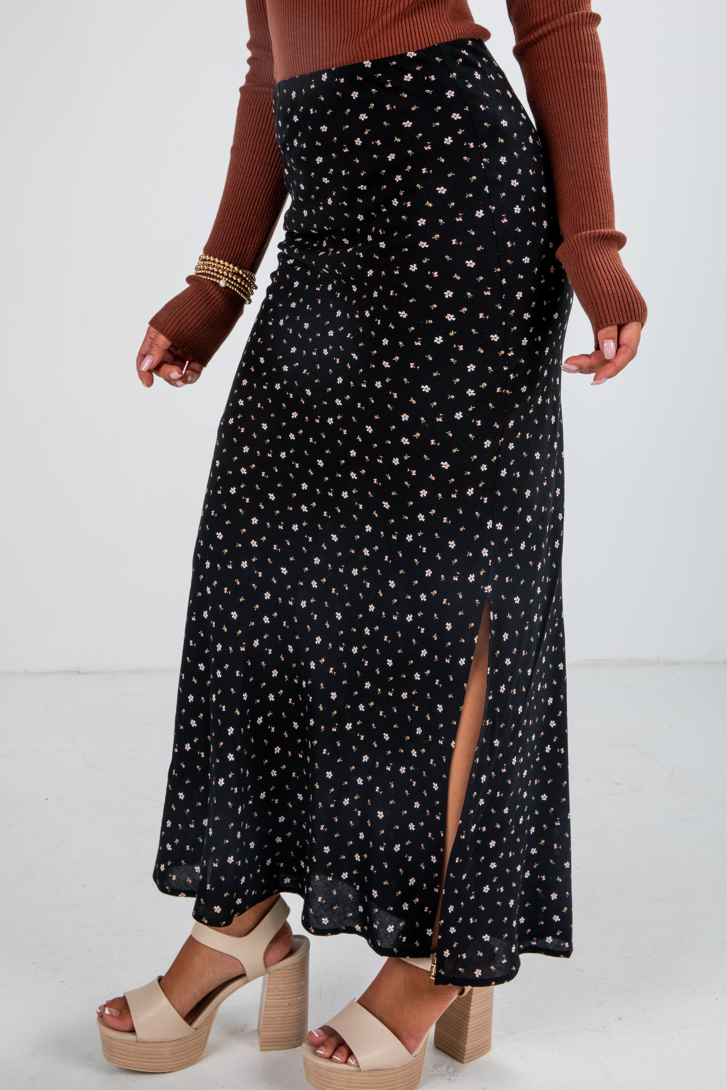 Person wearing a black patterned skirt with a brown sweater and beige sandals on a white background