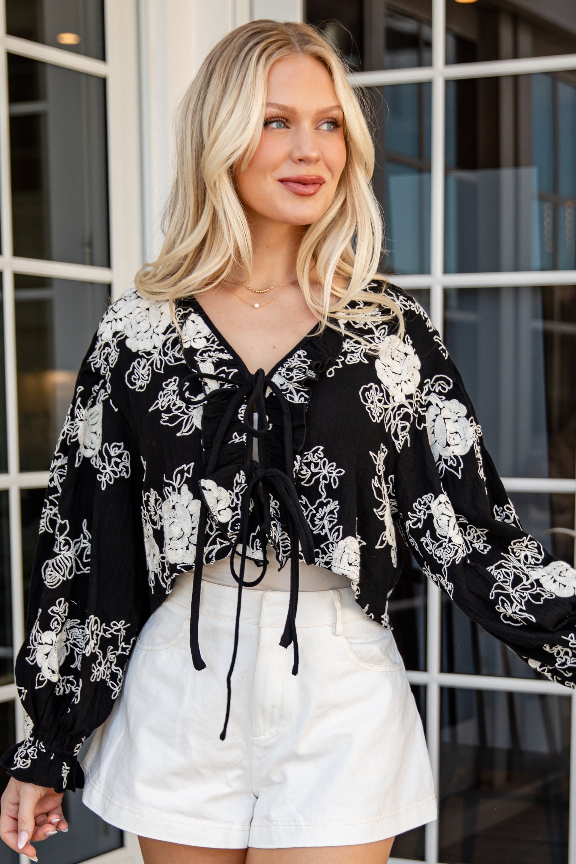 Woman wearing a black floral blouse and white shorts standing in front of glass doors.